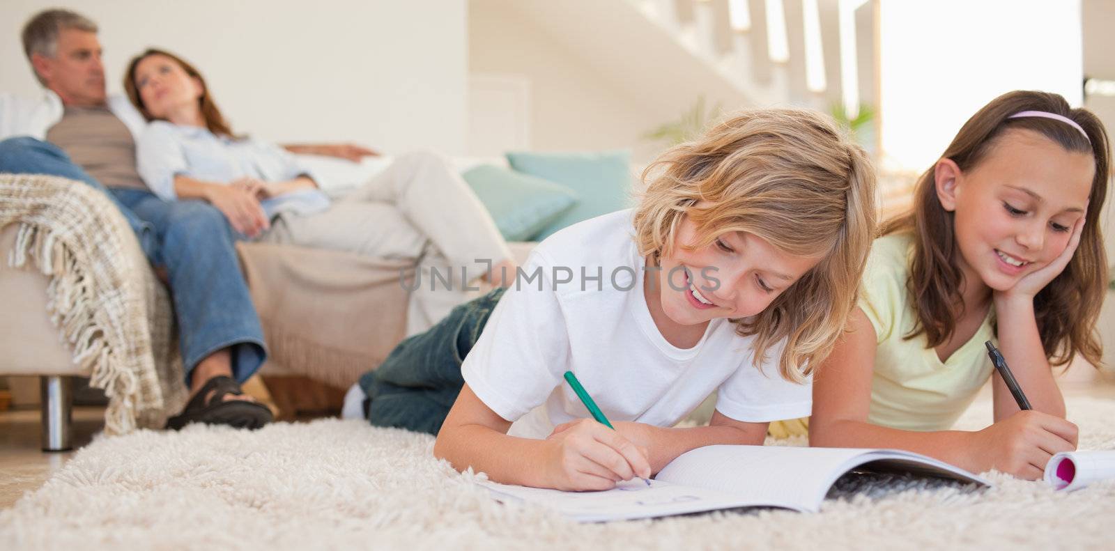 Siblings doing their homework on the carpet with parents behind  by Wavebreakmedia