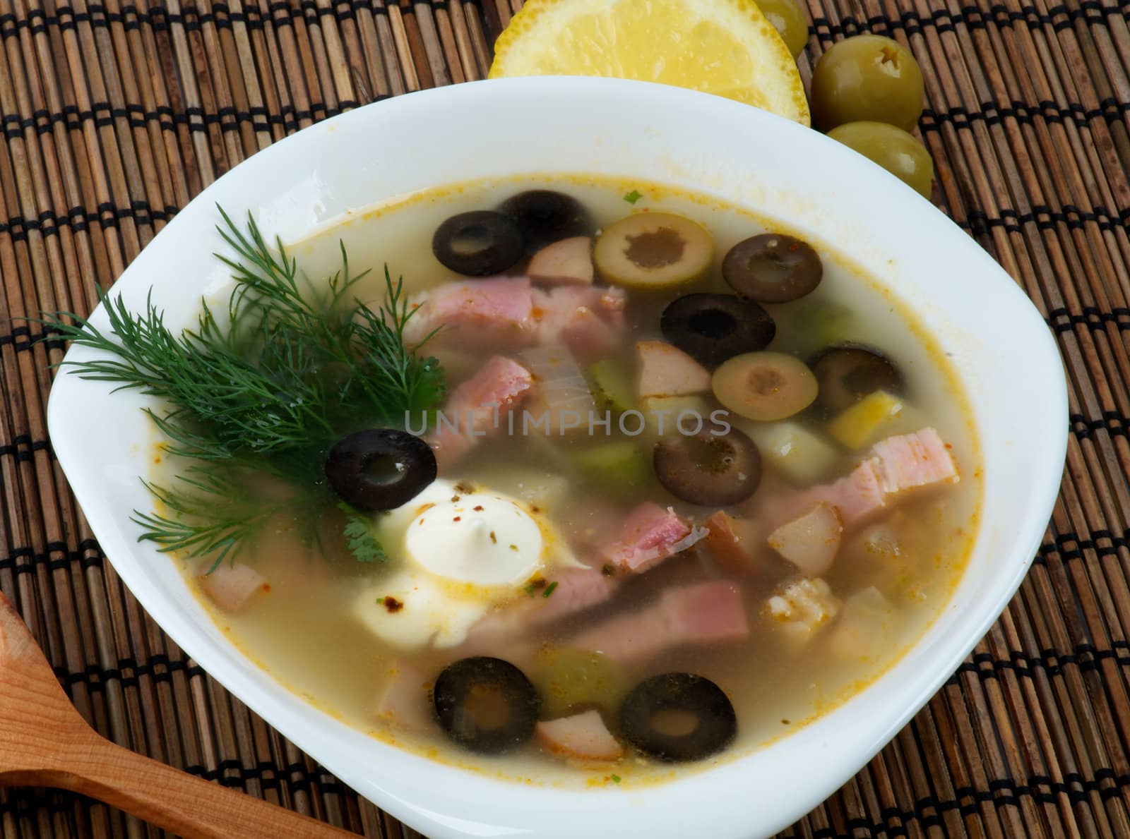 Russian Traditional Spicy Soup Solyanka Served with Lemon, Dill and Green Olives closeup on straw mat