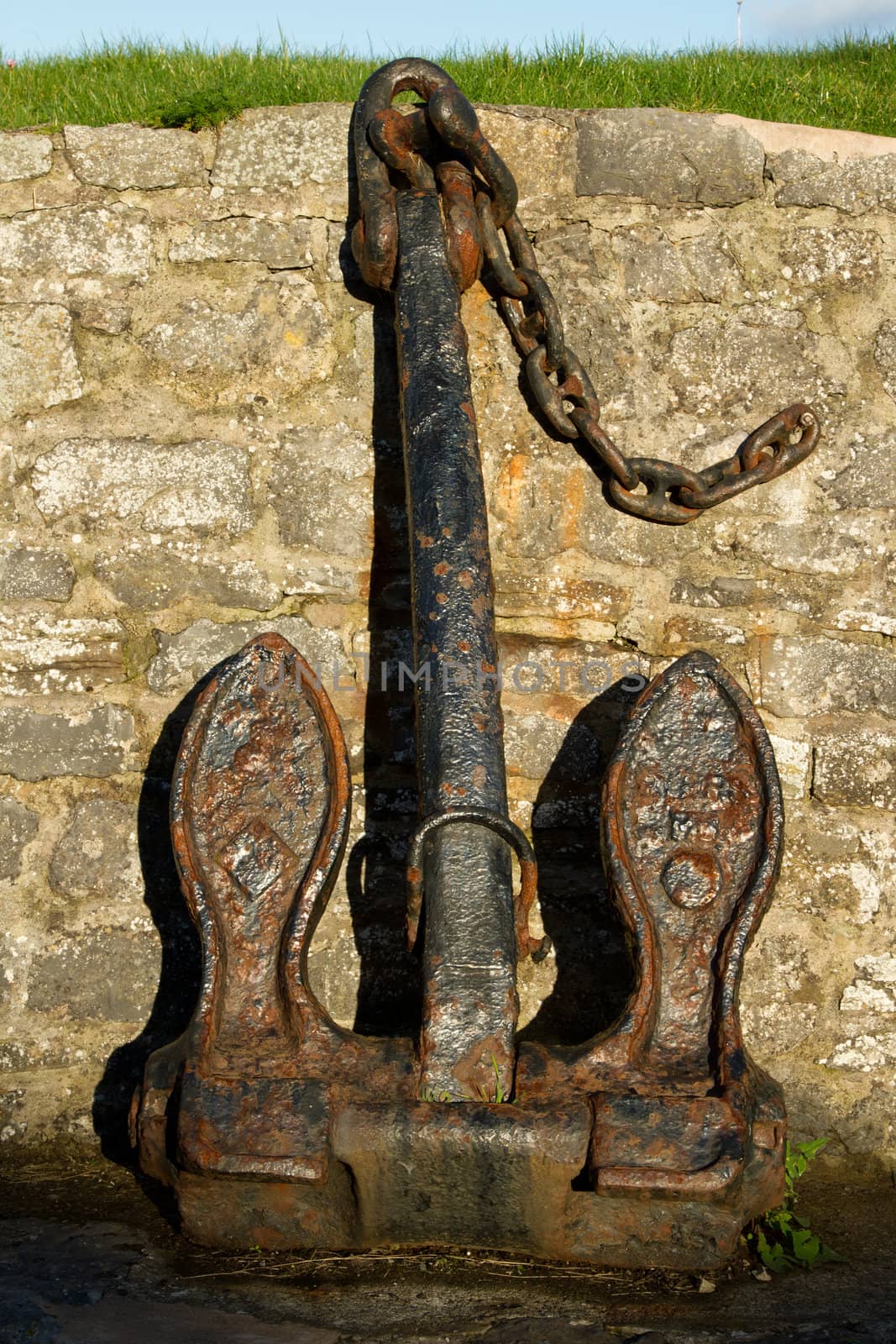 A vintage ship's anchor, rusted with black paint and chain, on display against a stone wall.