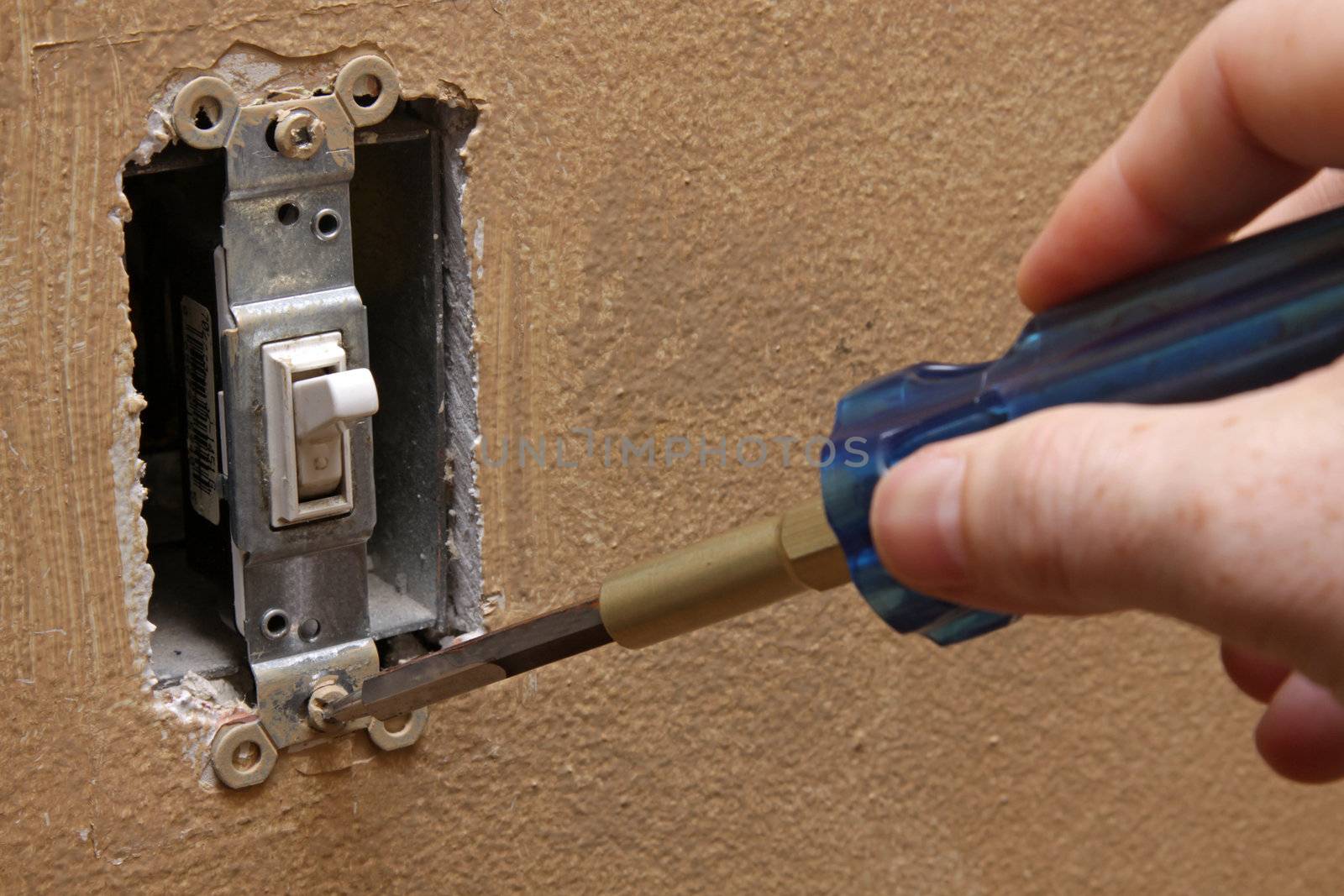 A hand using a screw driver to unscrew a screw holding a light switch.