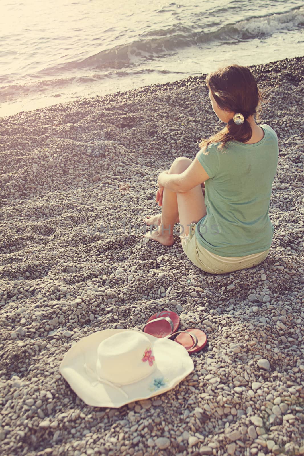The girl is sitting on the beach and watching the sea, next to a hat and slippers