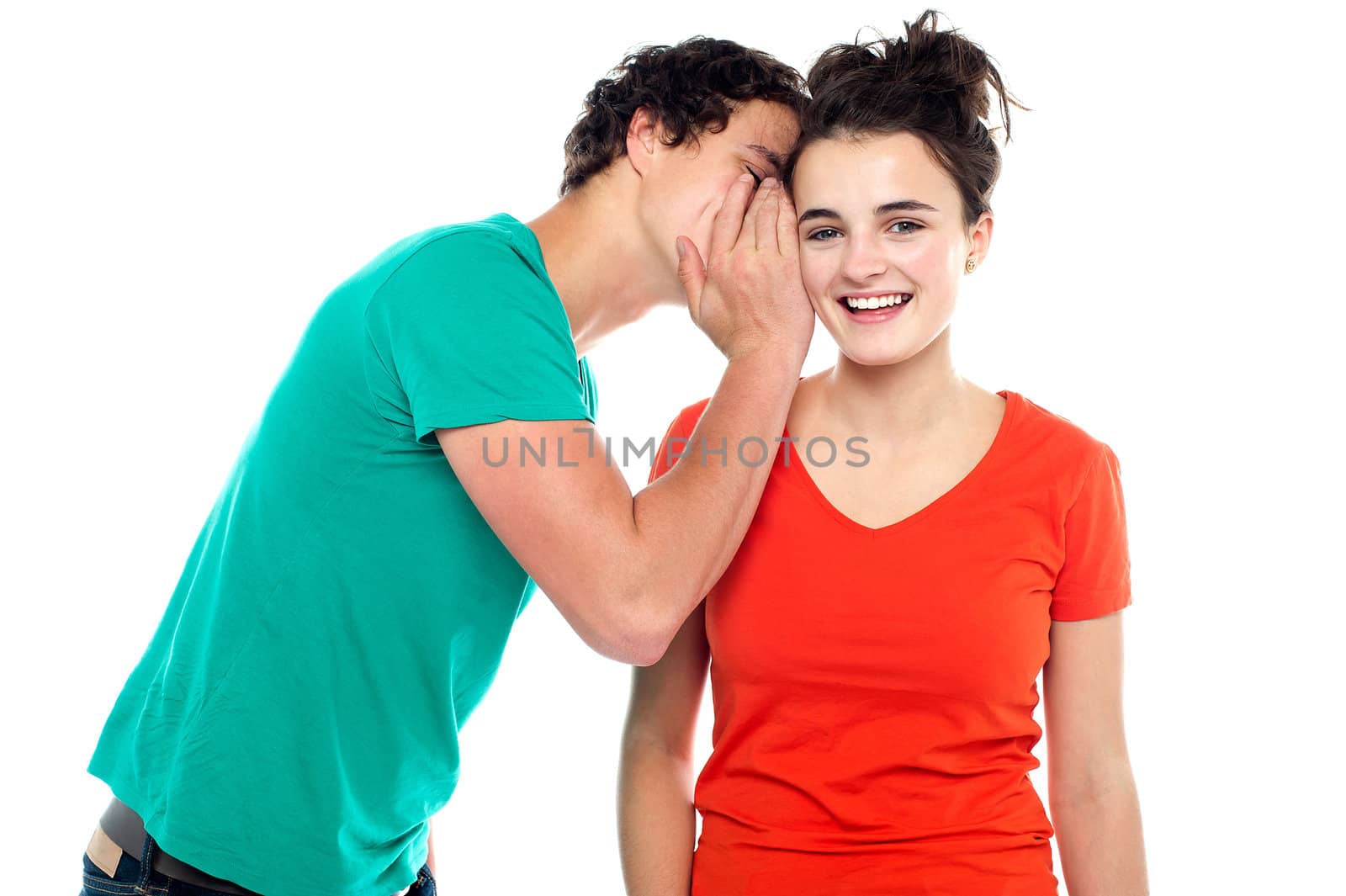 Handsome boy talking secret to young girl in her ear, girl laughing over isolated white background