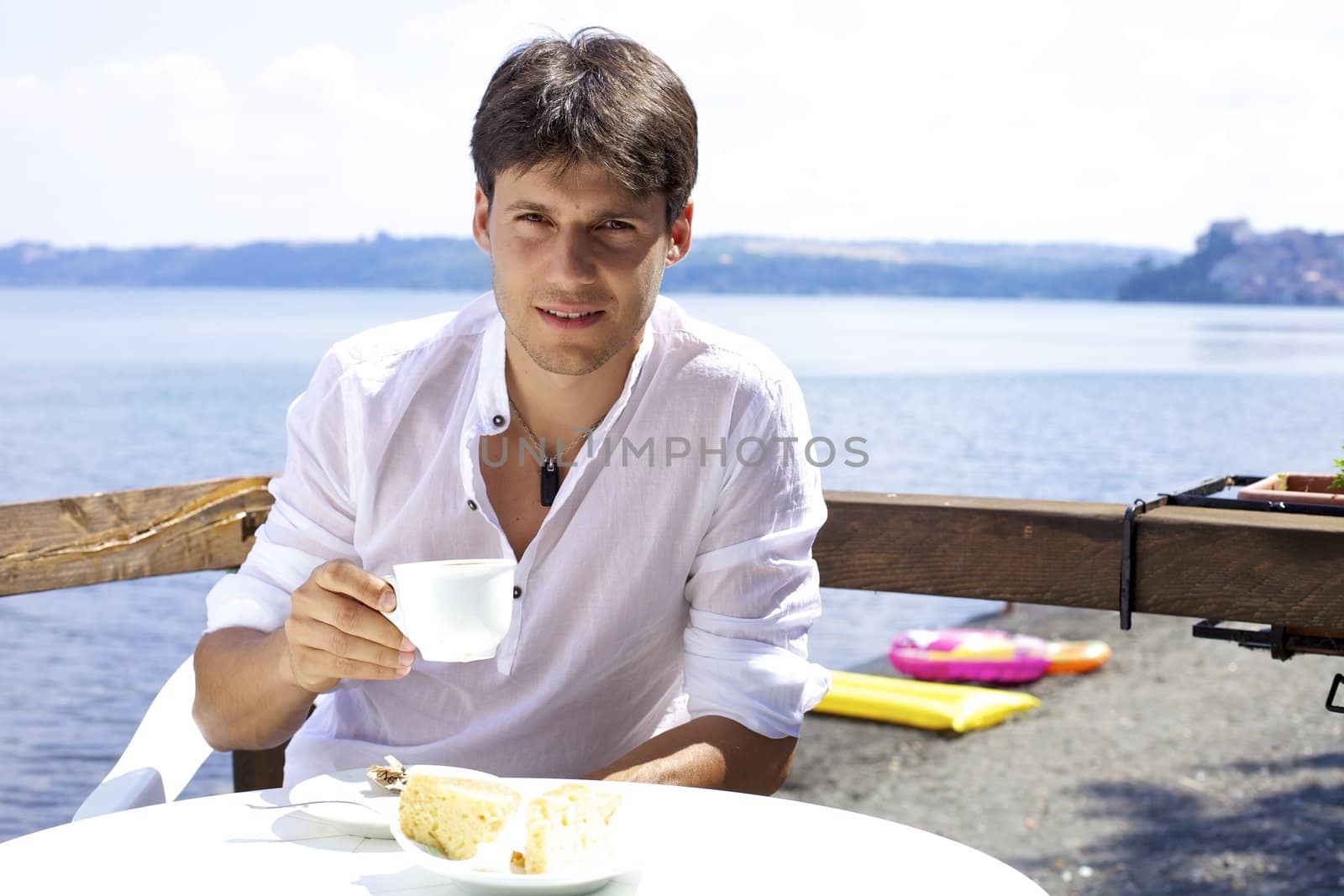 Good looking young man drinking coffee relaxed in front of lake smiling