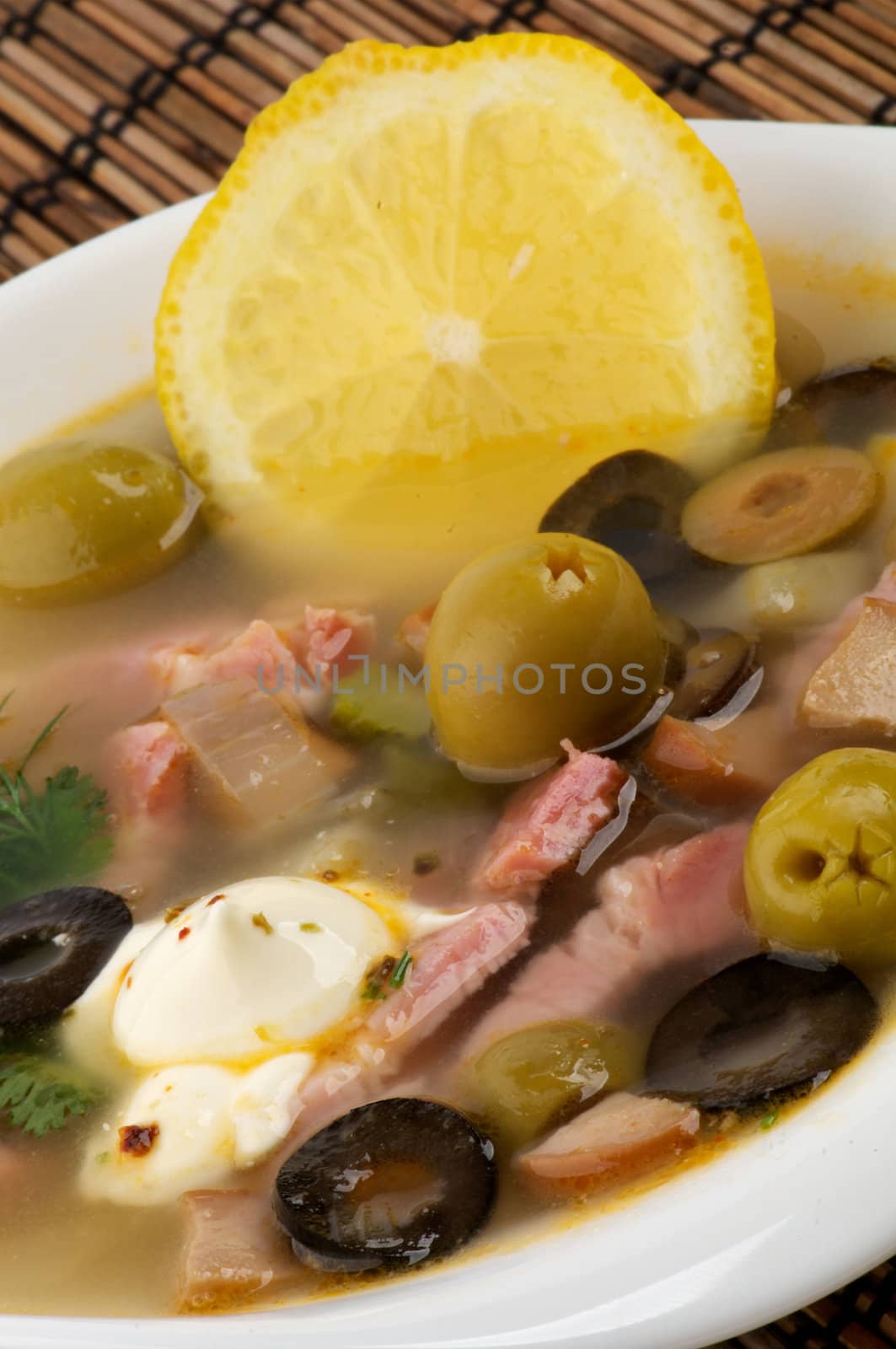 Russian Traditional Soup Solyanka with Lemon, Sour Cream and Green Olives in White Bowl closeup