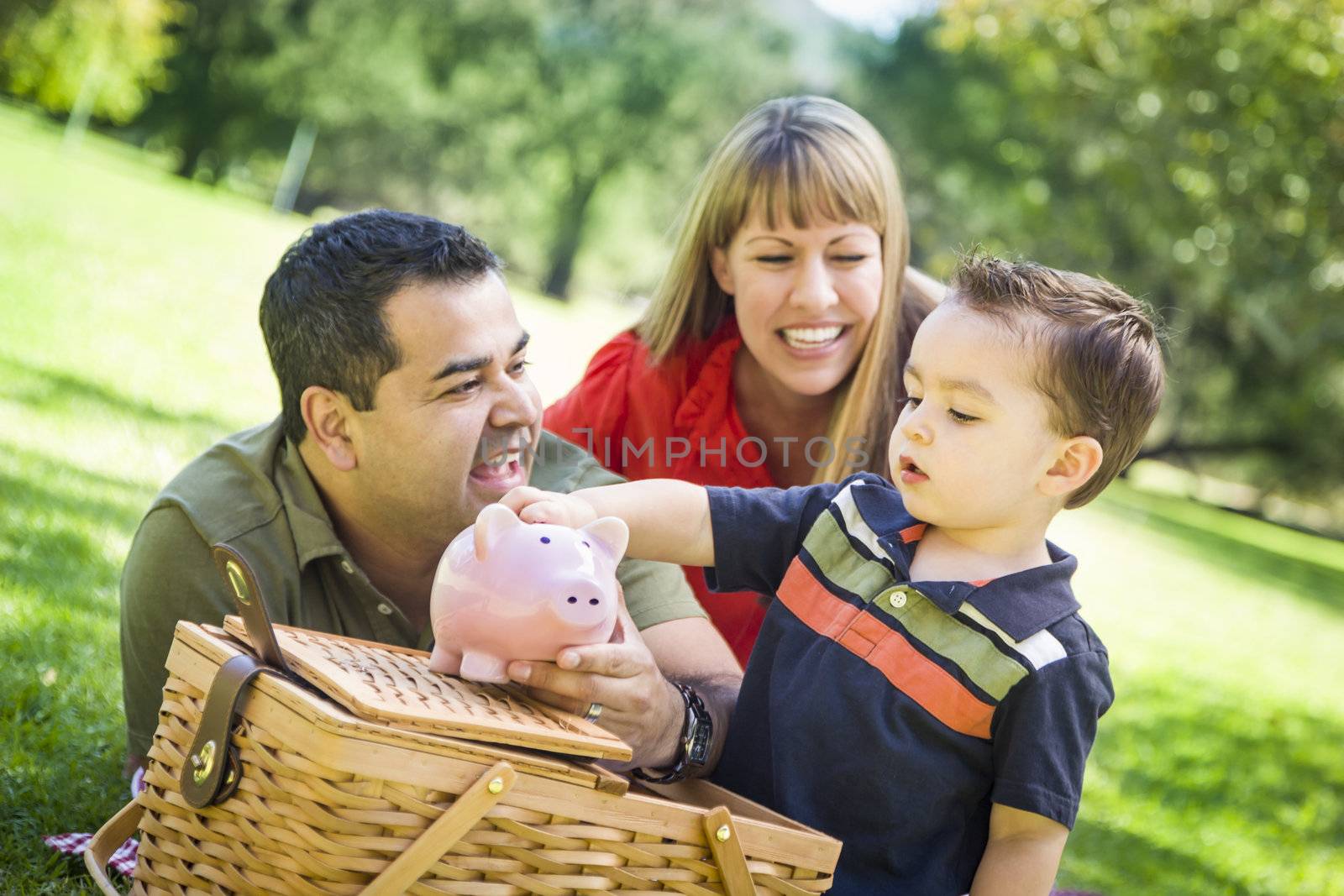 Happy Mixed Race Couple Give Their Son a Piggy Bank at a Picnic in the Park.