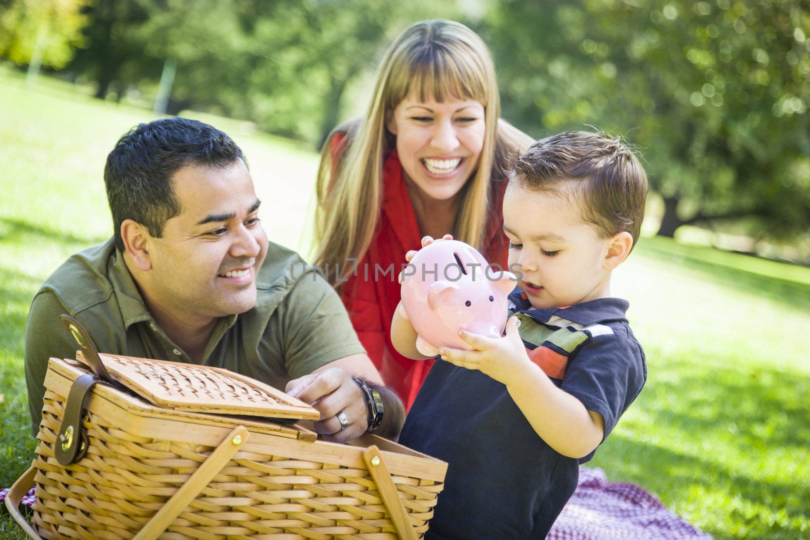 Happy Mixed Race Couple Give Their Son a Piggy Bank at a Picnic in the Park.