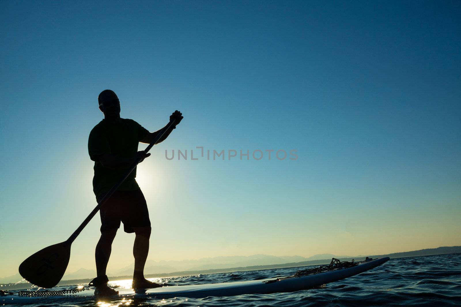 Man paddling stand up paddle board at sunset with blue sky in background.