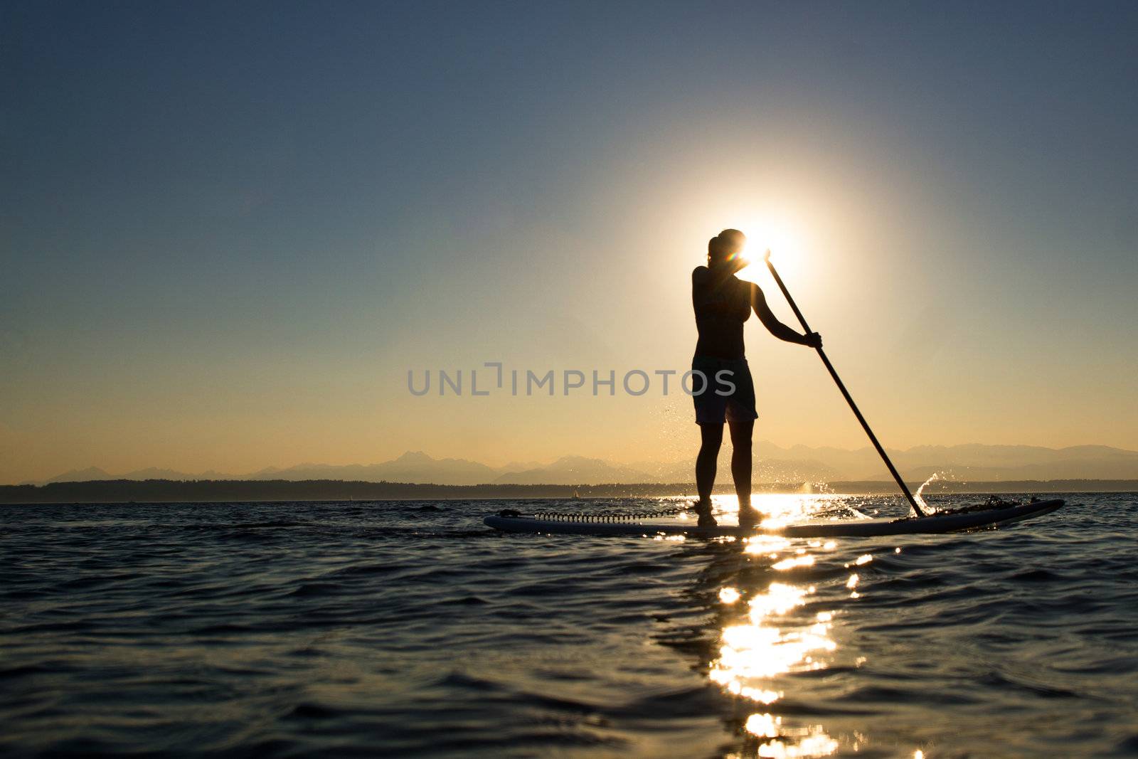 Woman paddling stand up paddle board at sunset with mountains in background.