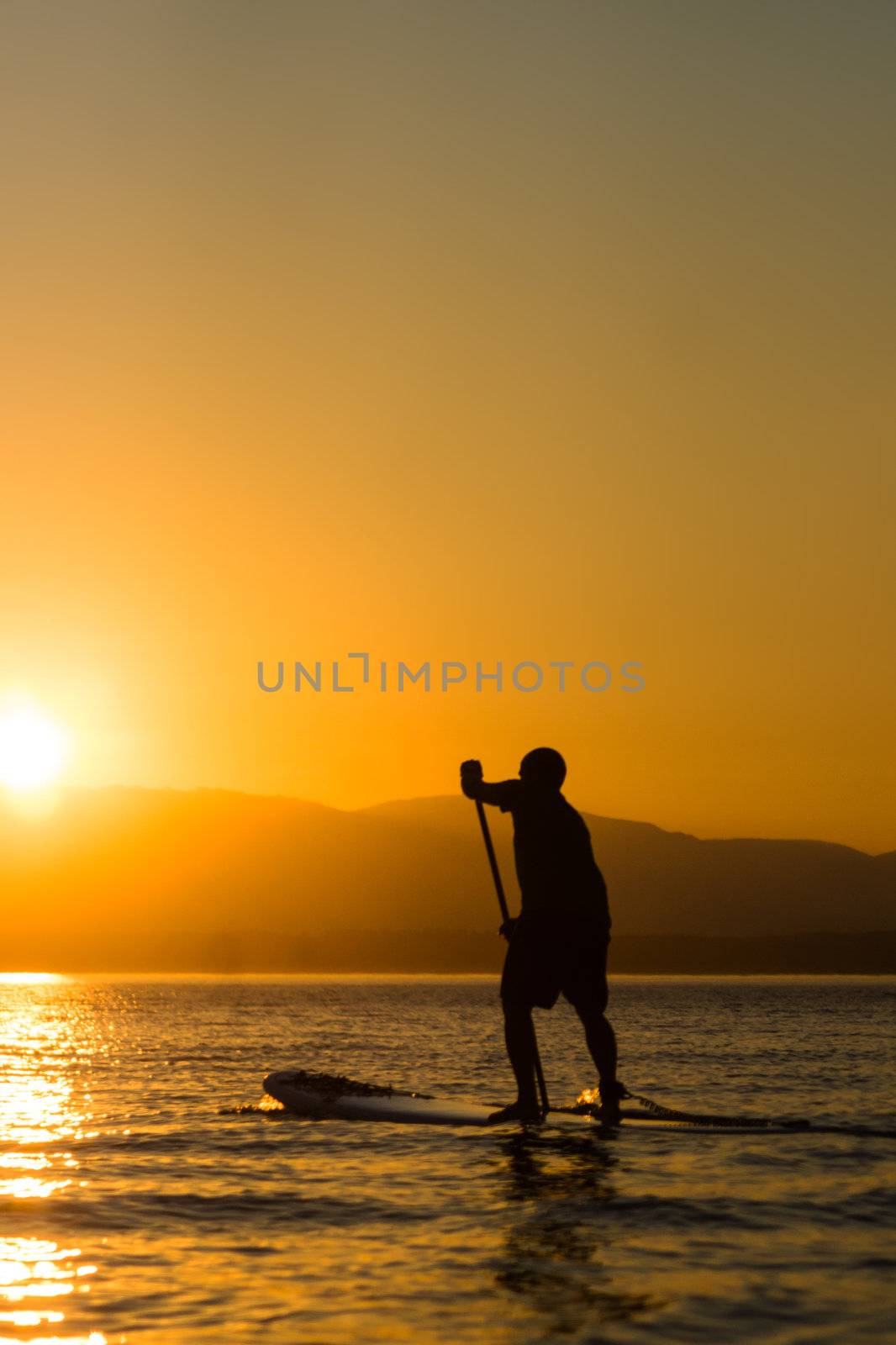 Man paddling stand up paddle board at sunset with mountains in background.