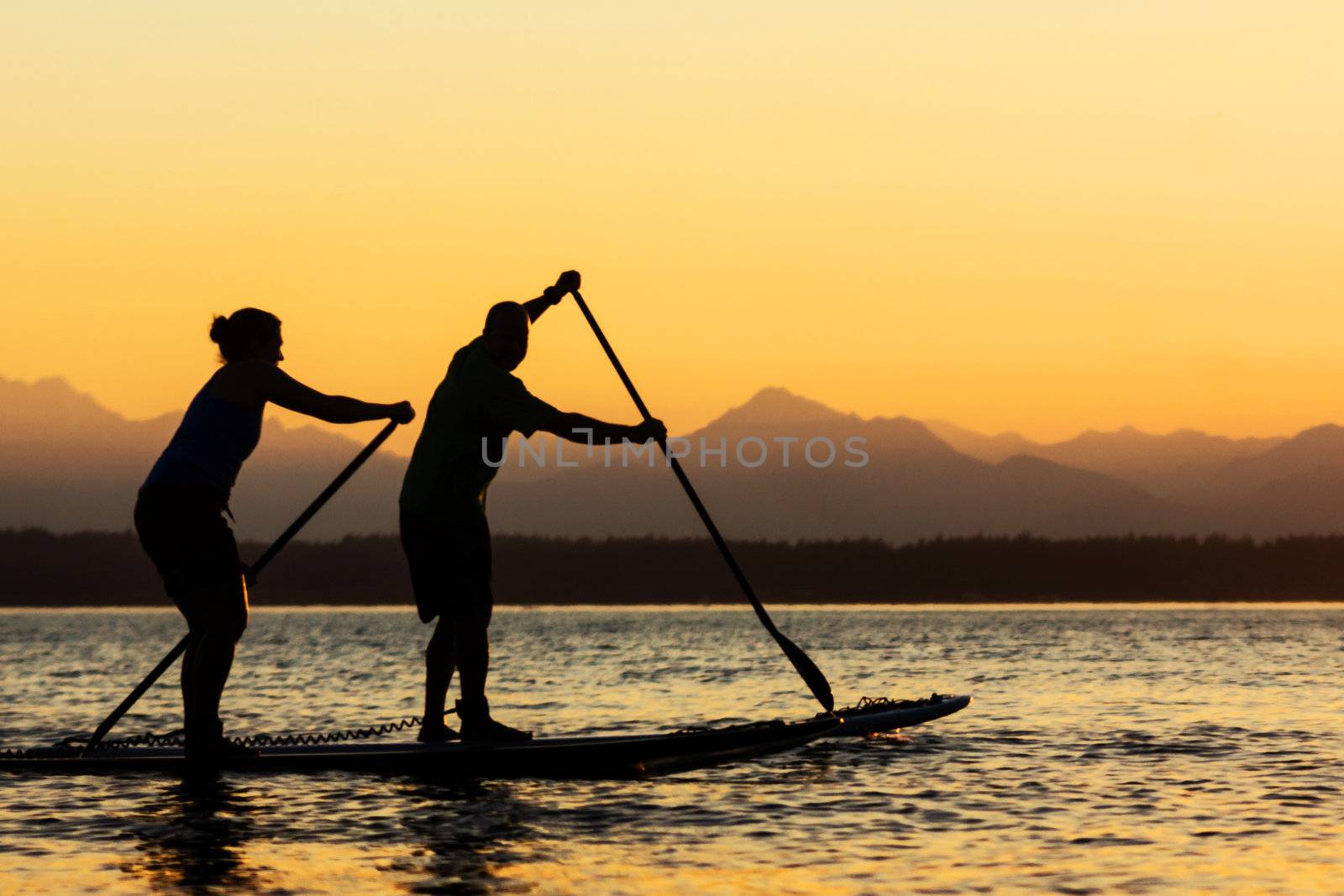 Couple paddling stand up paddle boards at sunset with mountains in background.