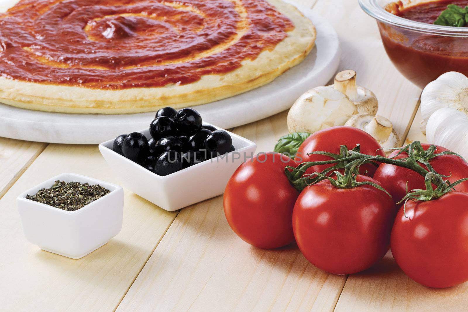 Tomato paste spread on raw pizza dough with tomato, mushrooms, black olives and crushed leaves on the side