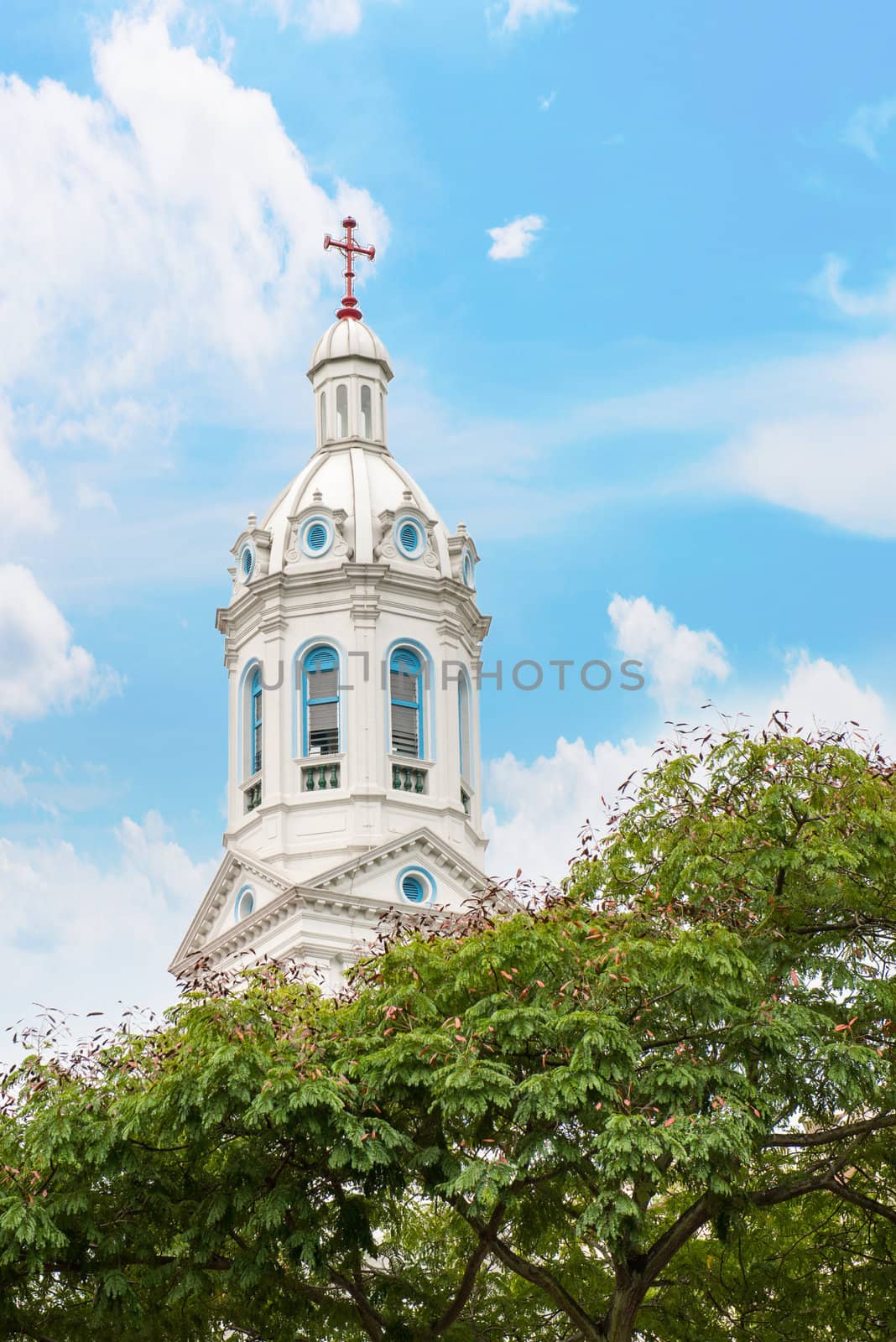 White church spire with trees and cloudy blue sky.Church of the Sacred Heart in Singapore.