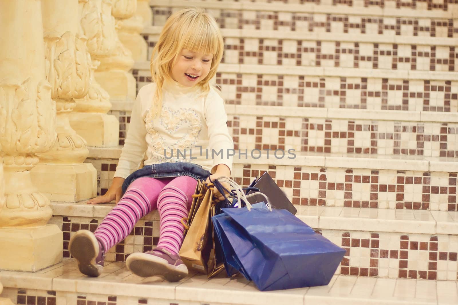 Smiling blond girl 3 years old in bright clothes sitting on the stairs with shopping