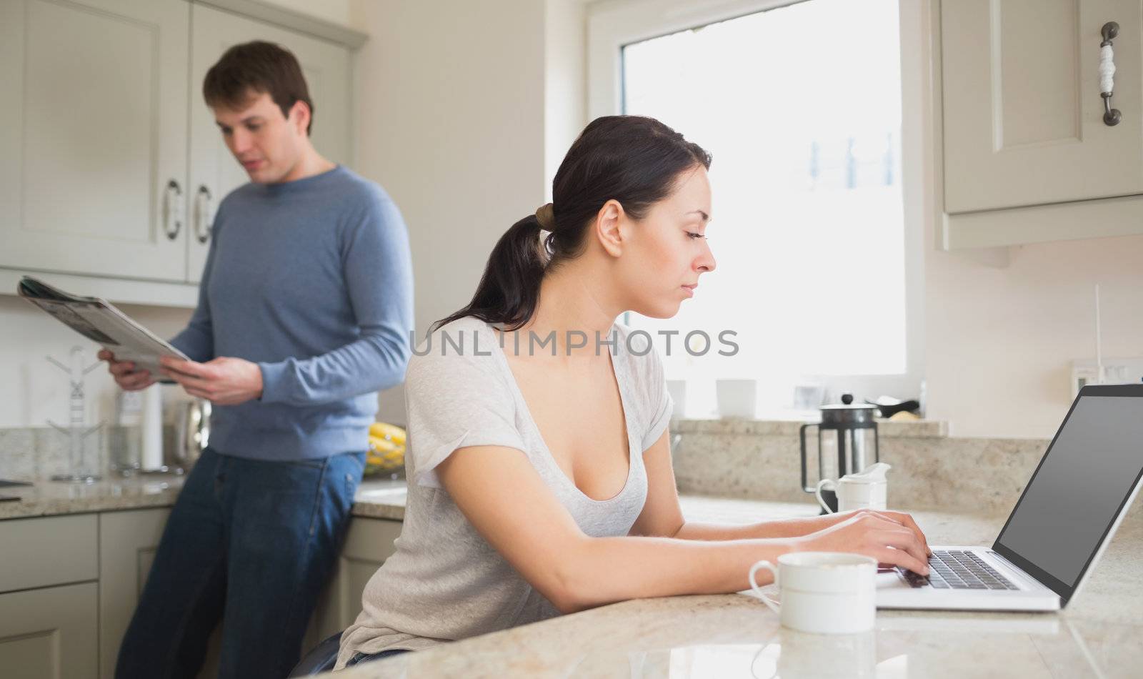 Two people in the kitchen who are using the laptop and reading a magazine