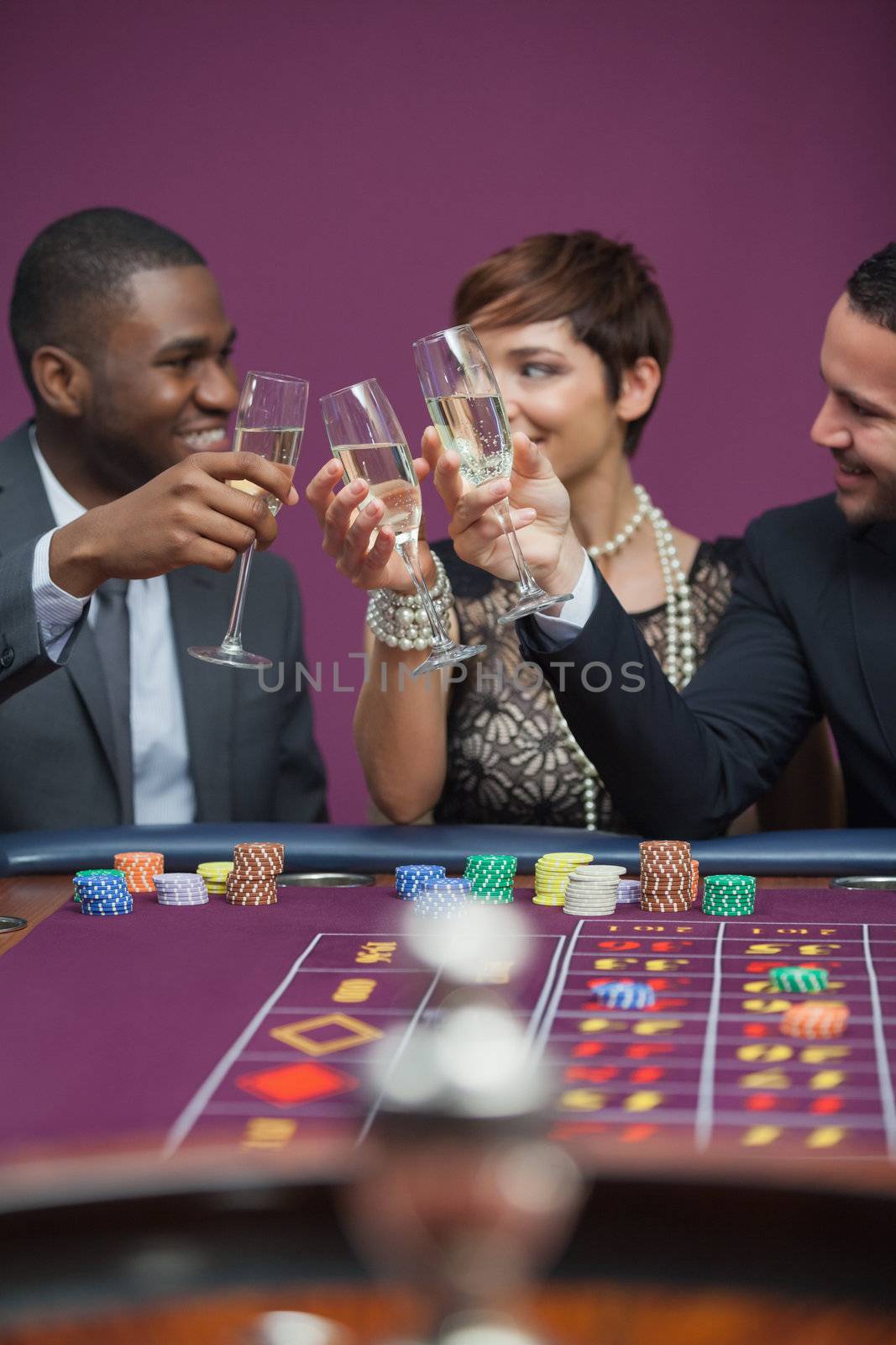 Three people playing roulette and toasting while sitting at table in a casino