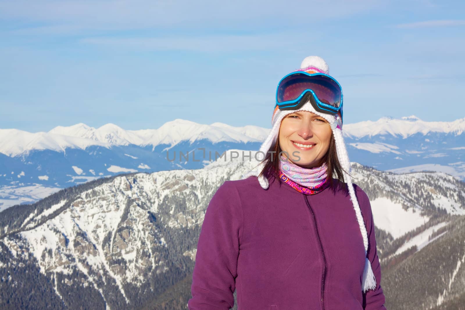 Smiling attractive young woman wearing knitted winter cap and ski glasses with picturesque winter mountains in the background