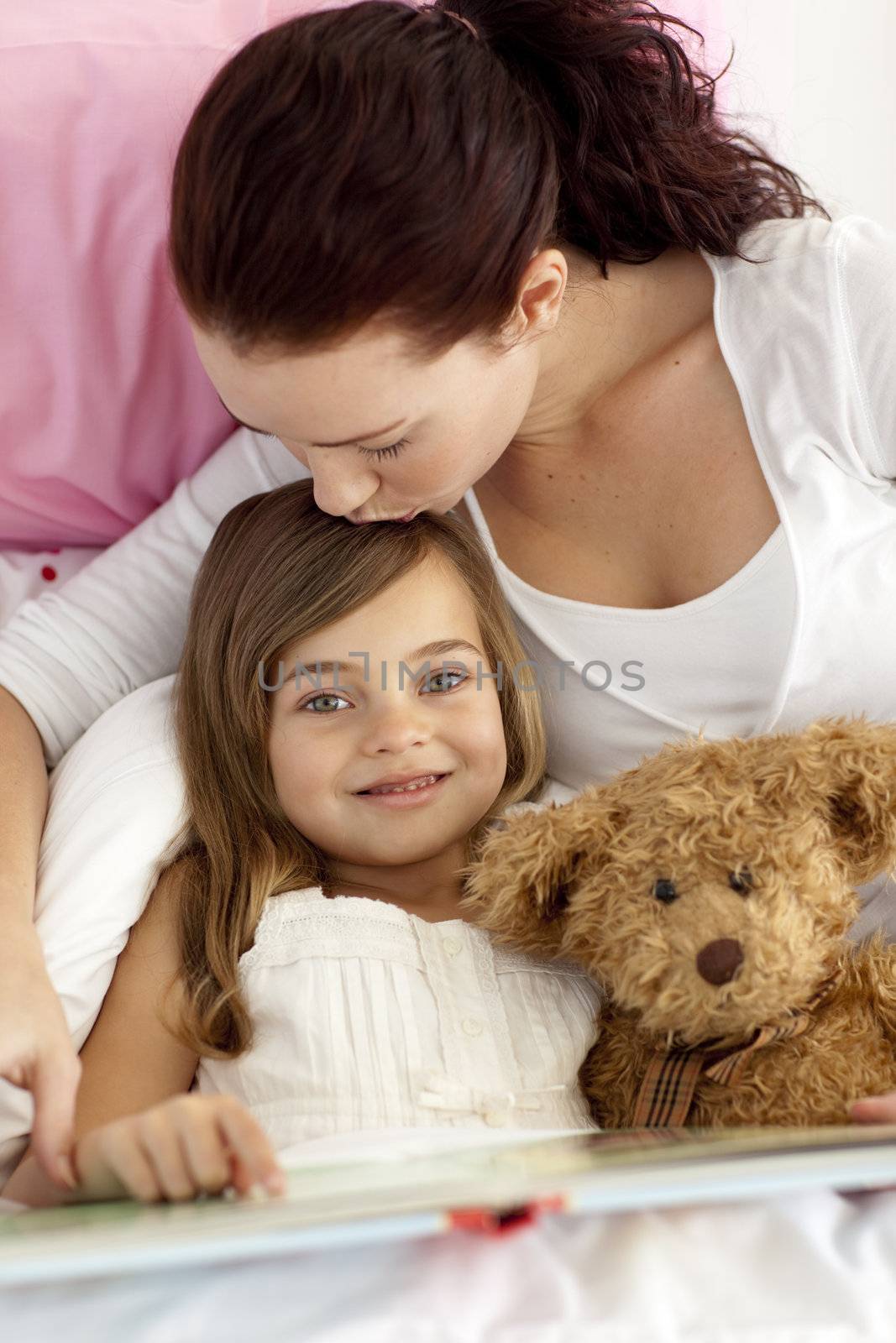 Mother kissing her daughter reading a book  in bed