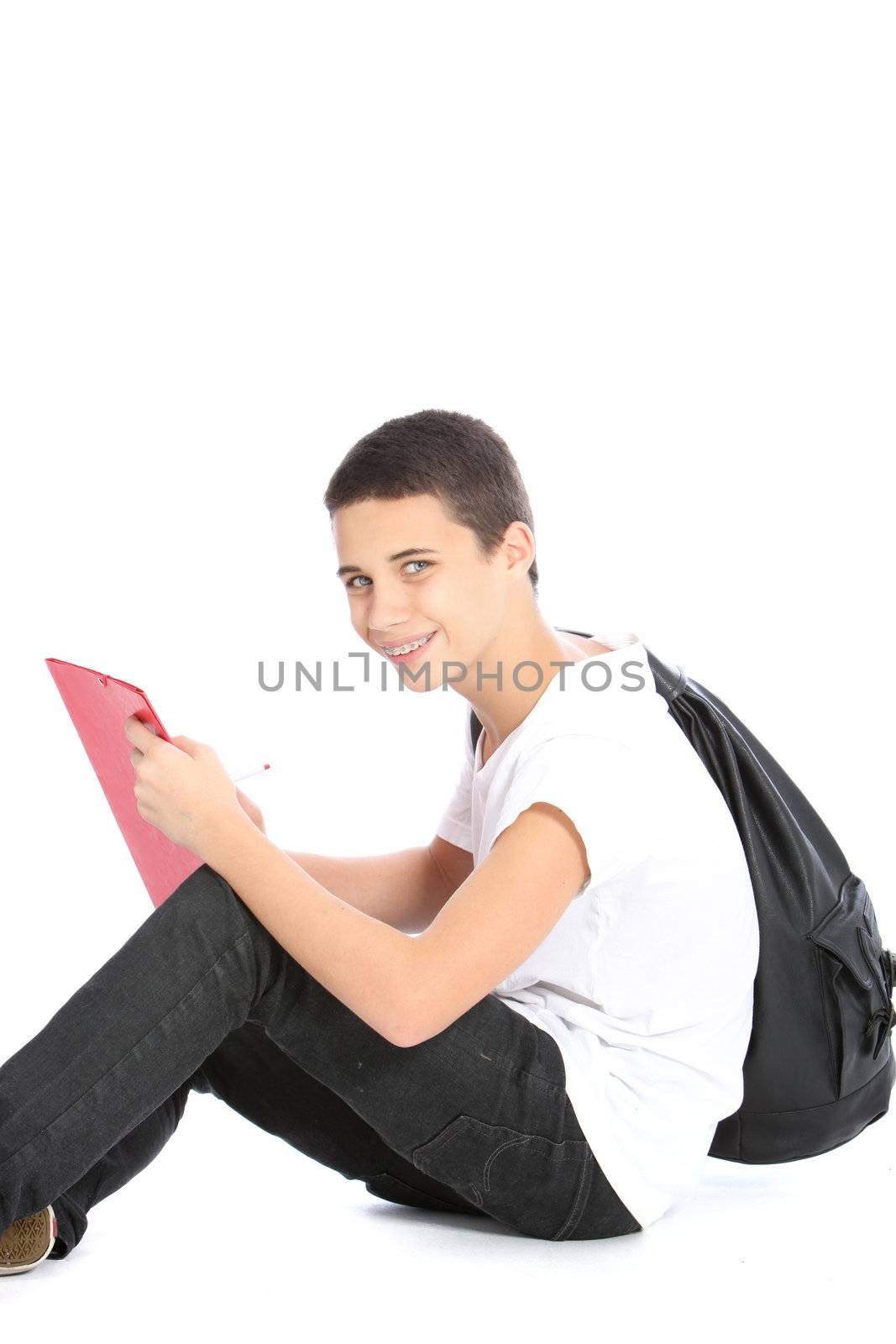 Young teenage boy scholar with a kit bag over his shoulder sitting on the floor writing in a folder isolated on white