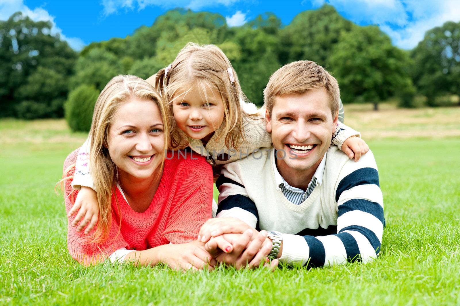 Cheerful couple lying on green grass with their sweet daughter child on top of them