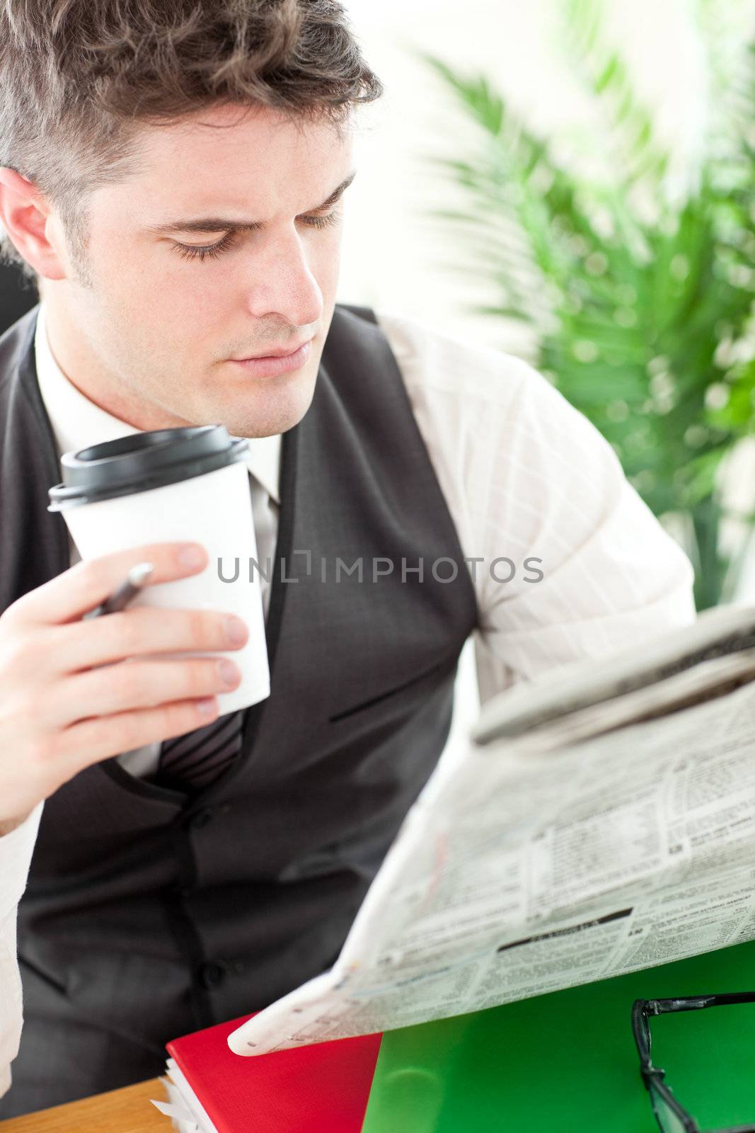 Charismatic businessman drinking a coffee while reading a newspaper sitting at his desk