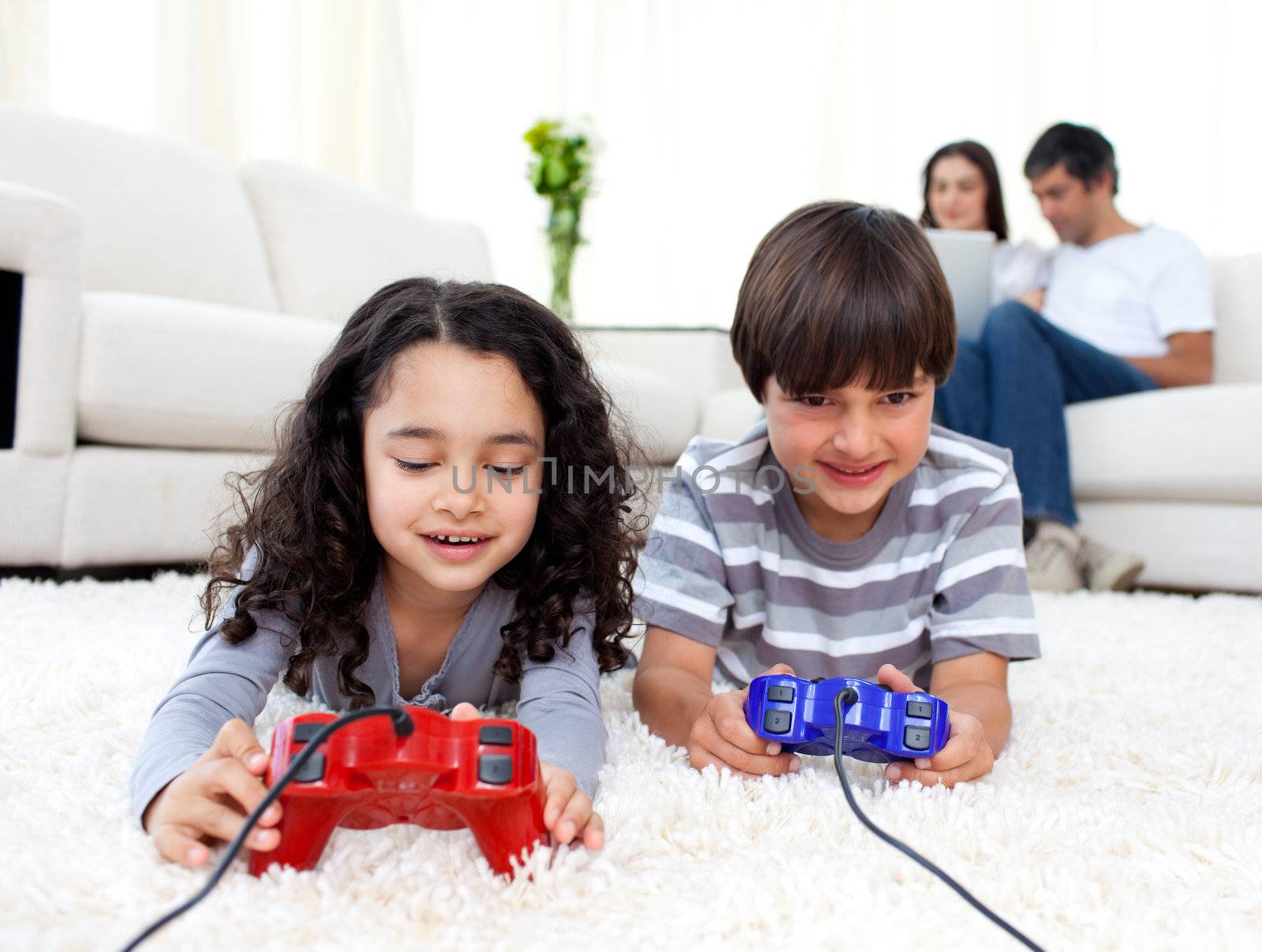 Jolly siblings playing video games lying on the floor with their parents in the background