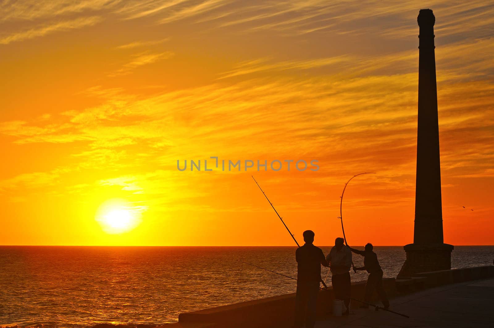 Silhouettes of three men on the coast fishing at sunset.