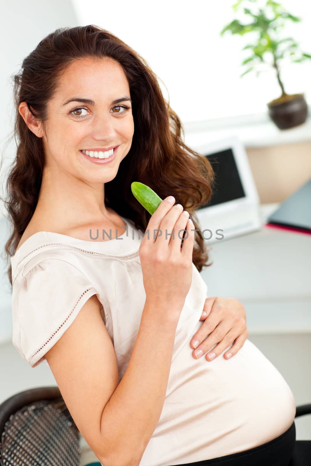 Bright pregnant woman eating a gherkin in her office sitting at her desk