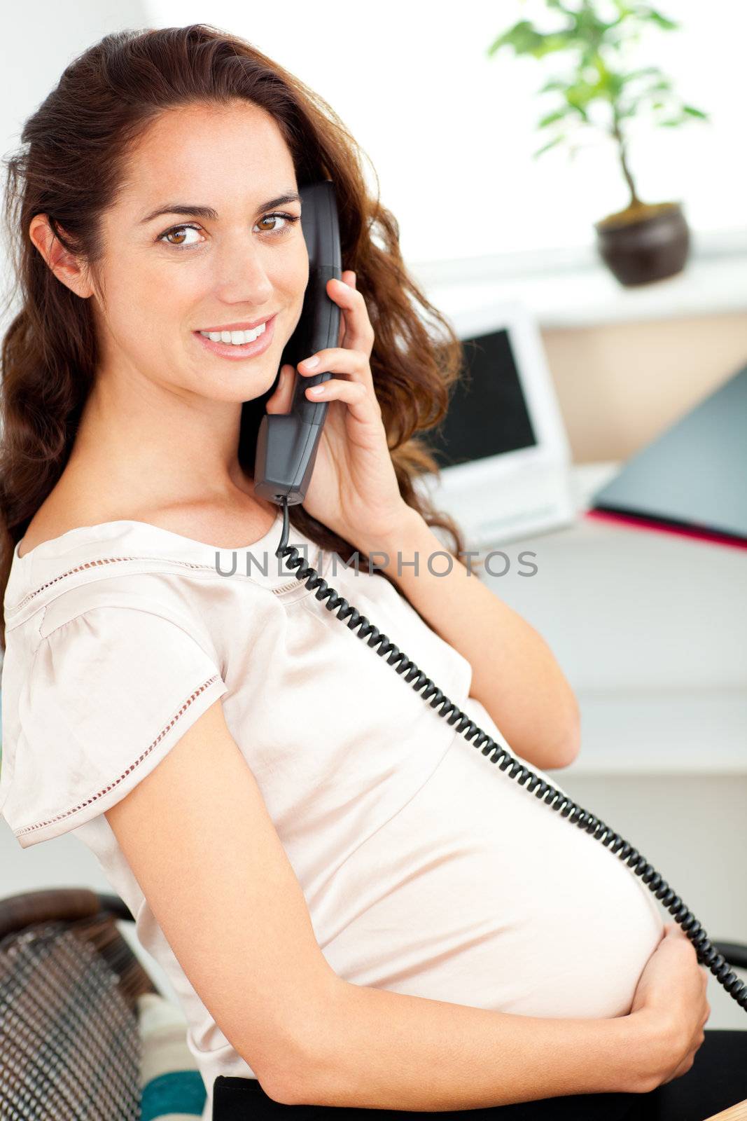 Charming pregnant businesswoman talking on phone sitting at her desk in her office