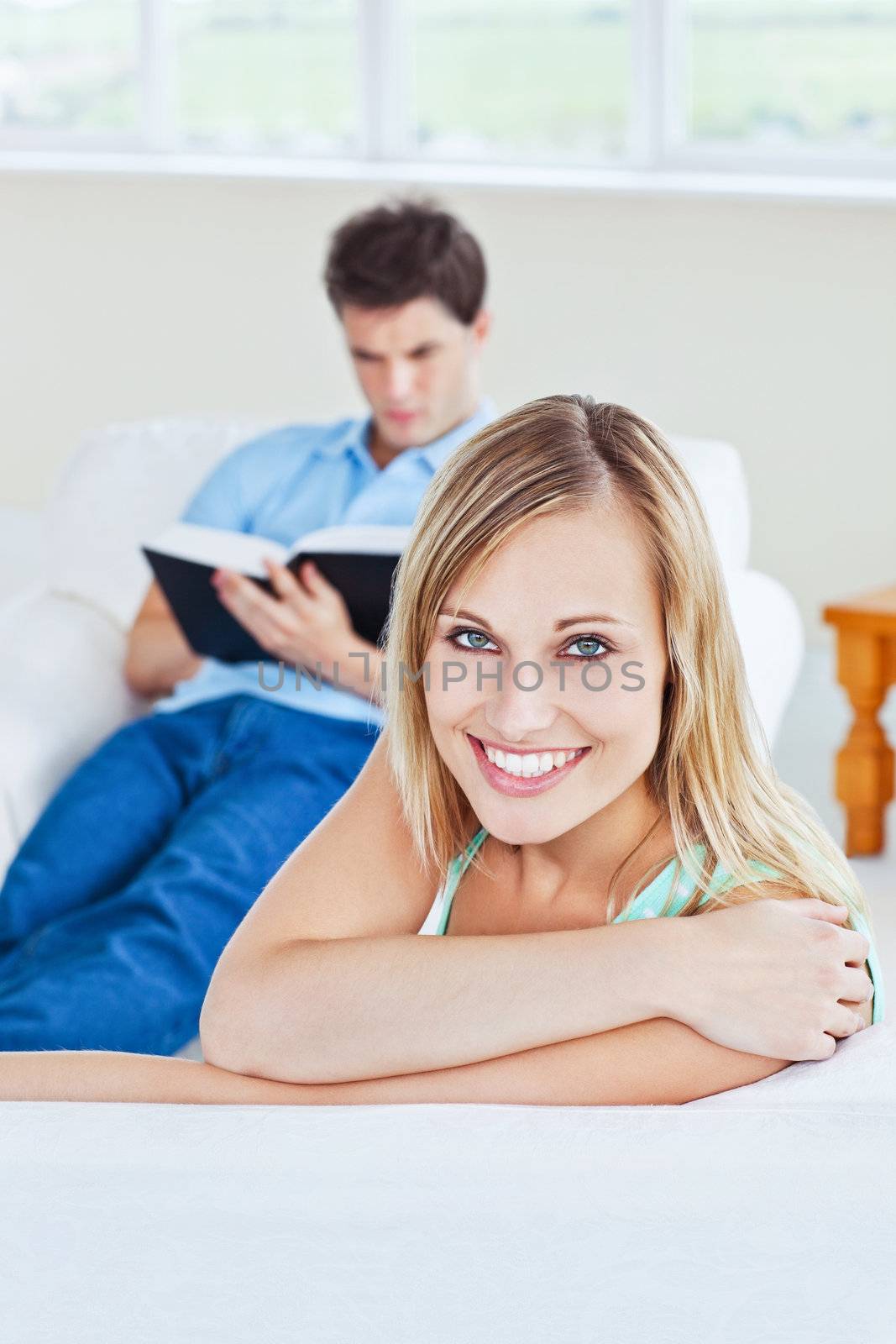 Girlfriend smiling at the camera while her boyfriend is focused on a  book sitting in the background