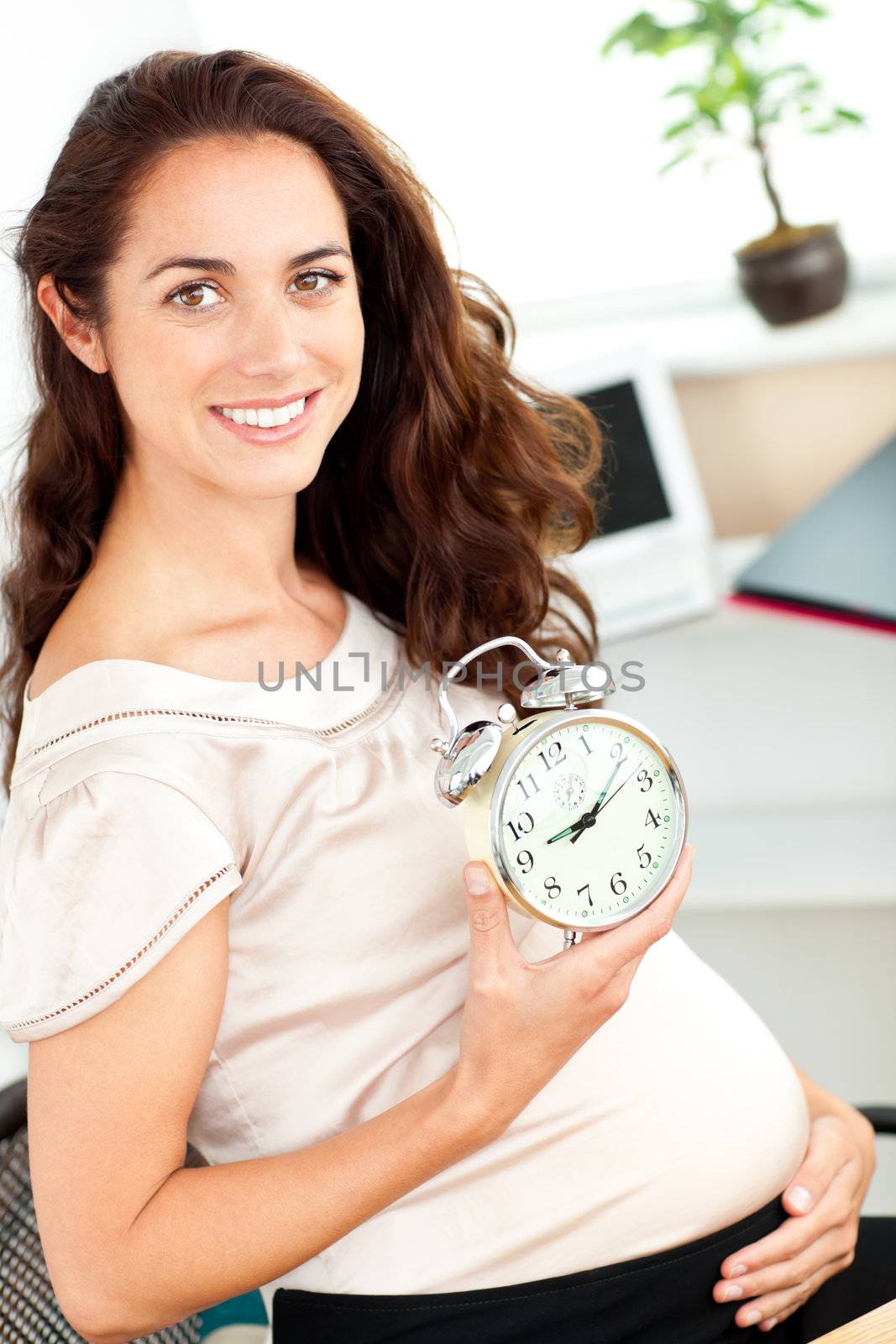 Radiant pregnant businesswoman holding an alarm clock and sitting at her desk in her office