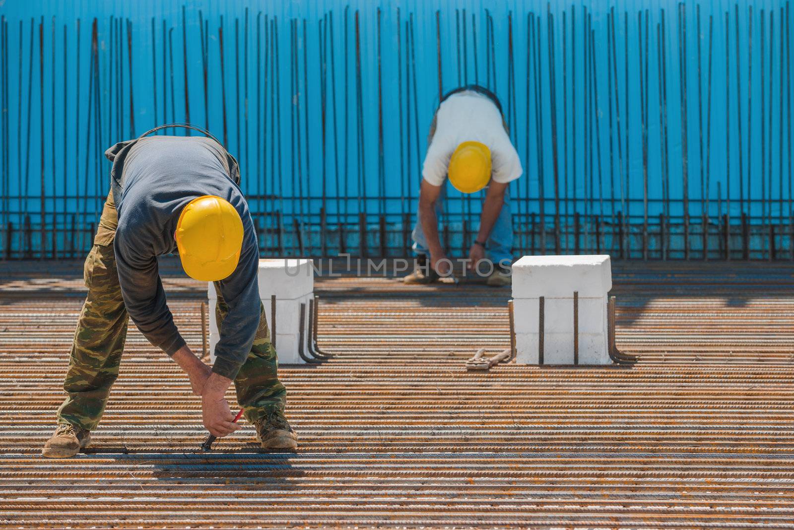 Authentic construction workers installing binding wires to reinforcement steel bars in front of a blue insulated surface prior to pouring concrete