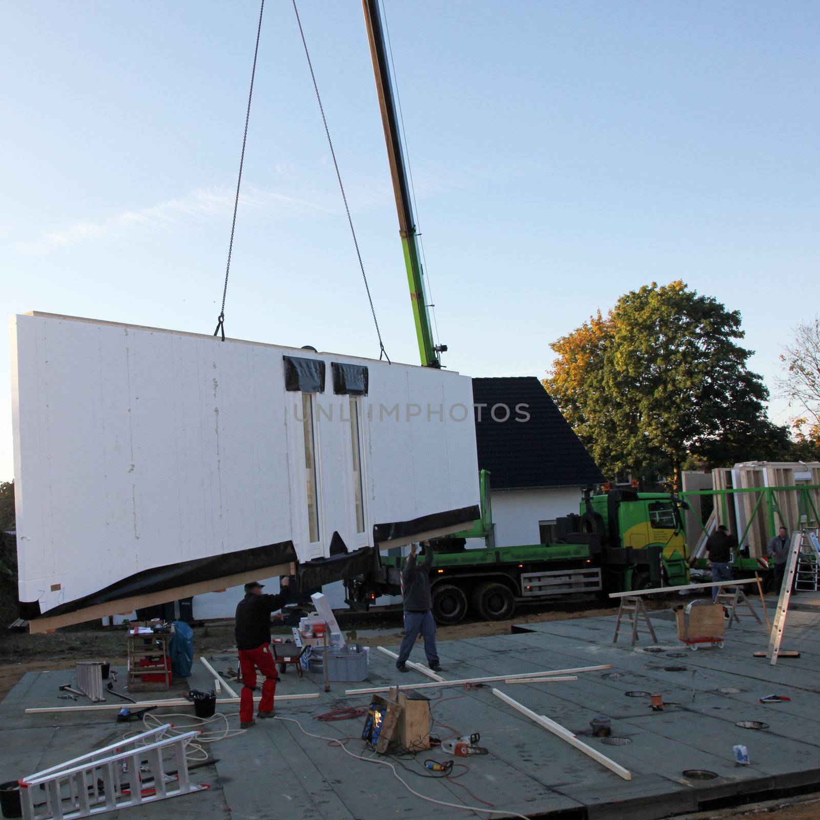 Workmen guiding in a prefabricated wall which is hanging suspended from a crane as it is delivered to a building site for installation onto the cement foundations