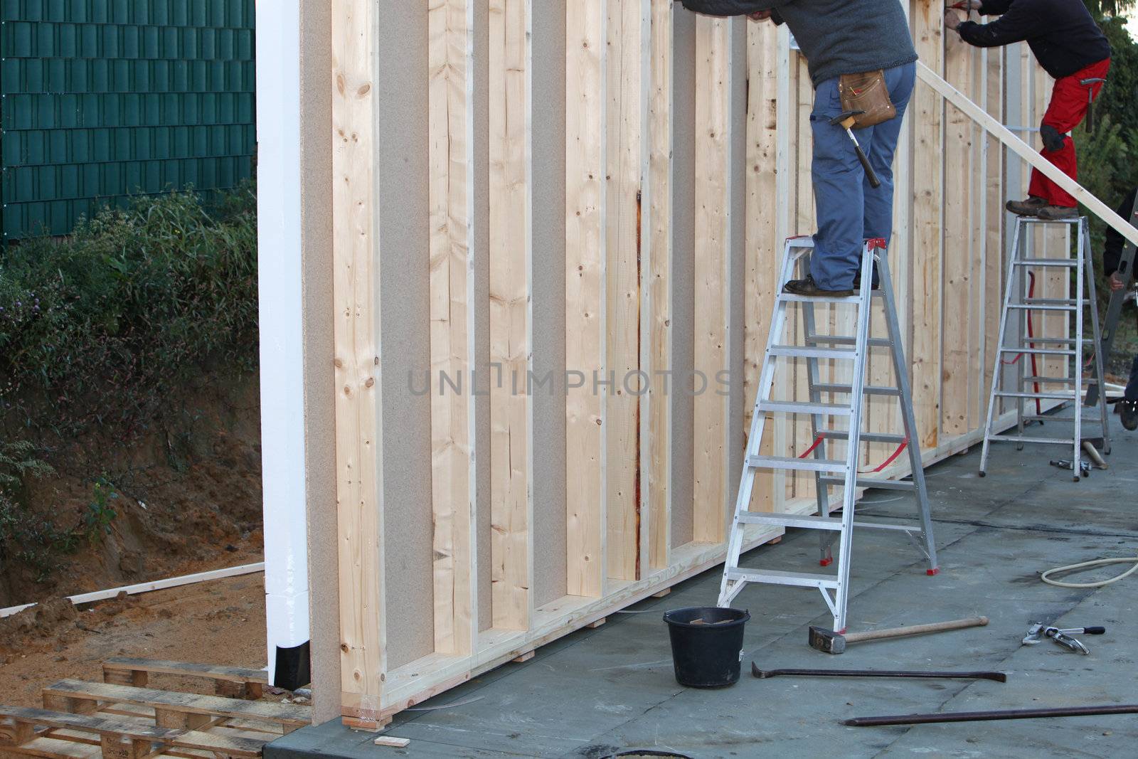 Builders standing on ladders installing a prefabricated exterior wall of a timber frame house on a cement foundation