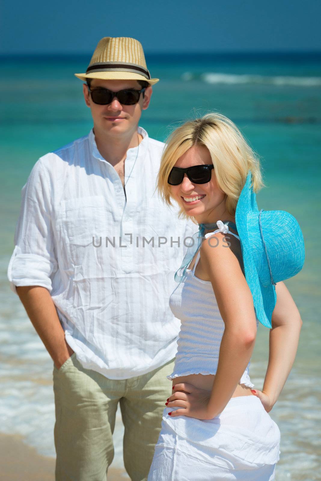 Happy young couple in sunglasses and white dress on a tropical beach with blue sea on background. Selective focus on the girl.