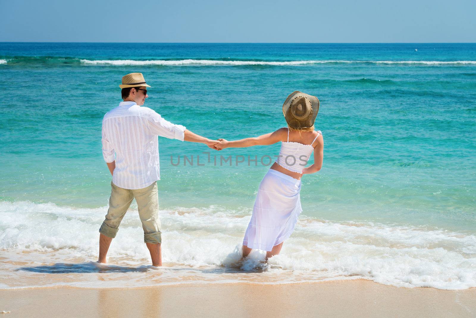 Rear view of romantic young couple in hats and white dress in blue sea at the beach 