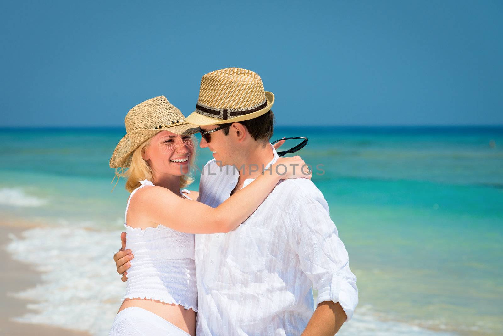Happy embracing young couple in hats and white dress on a tropical beach with blue sea on background