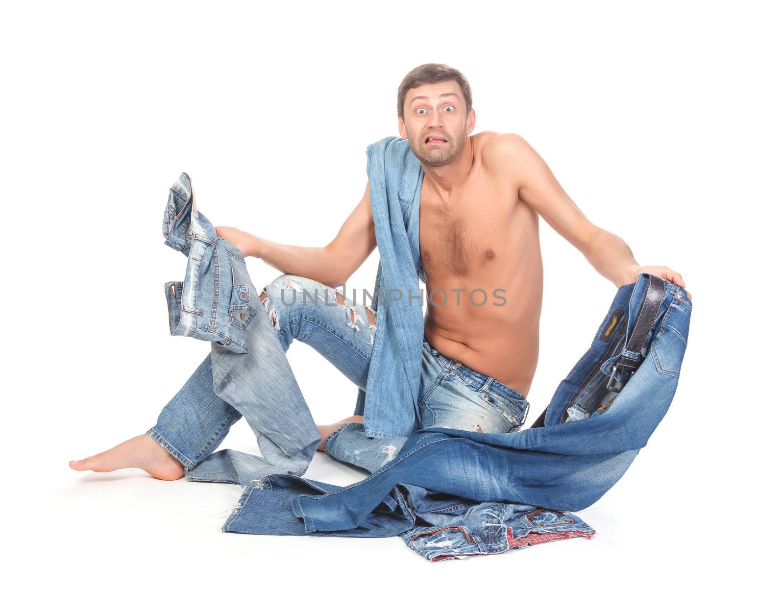 Attractive Indecisive man trying to dress sitting shirtless and barefoot on the floor holding several pairs of denim jeans shrugging his shoulders with a look of consternation