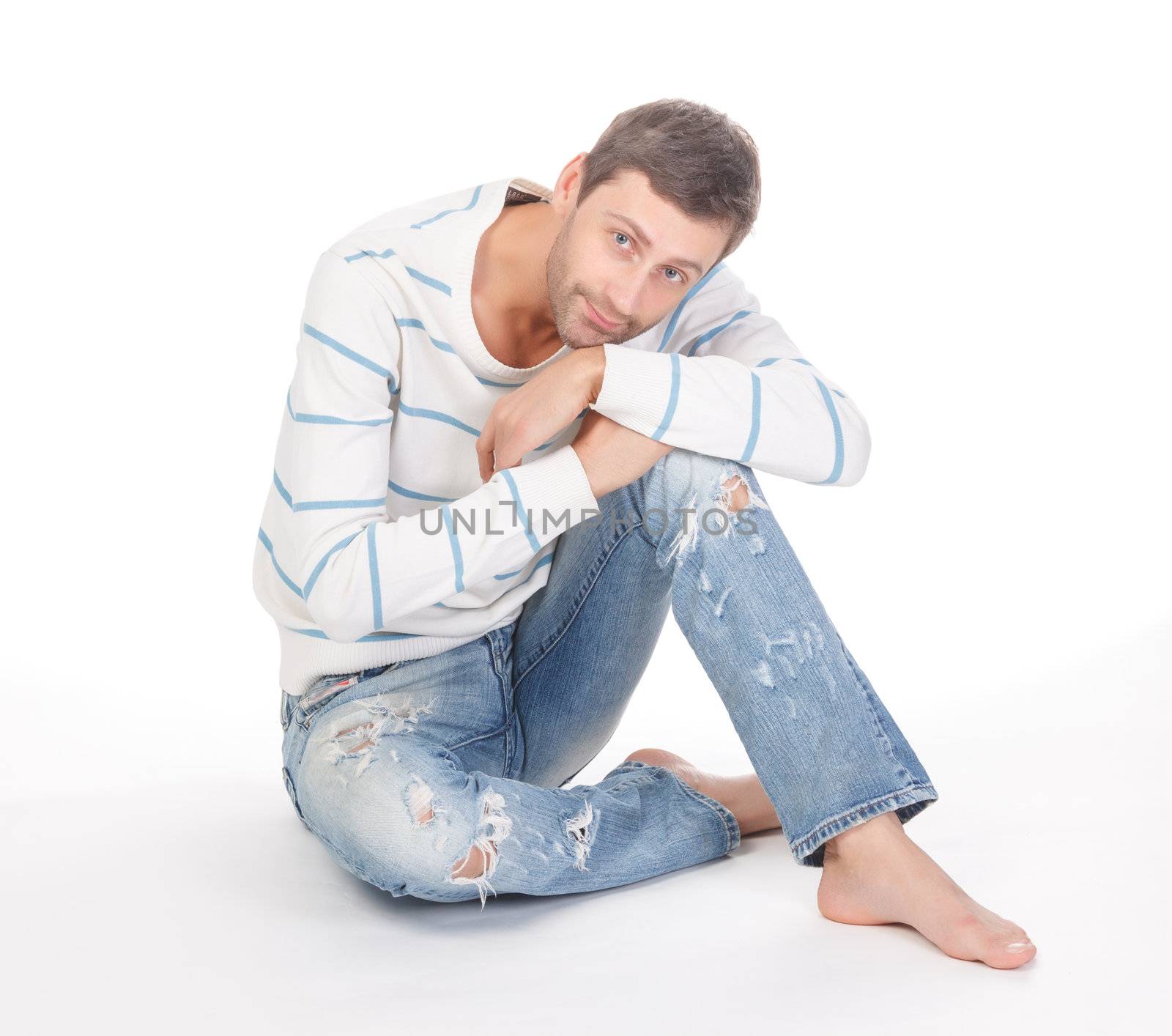 Handsome casual man sitting barefoot in trendy ragged jeans over a white background