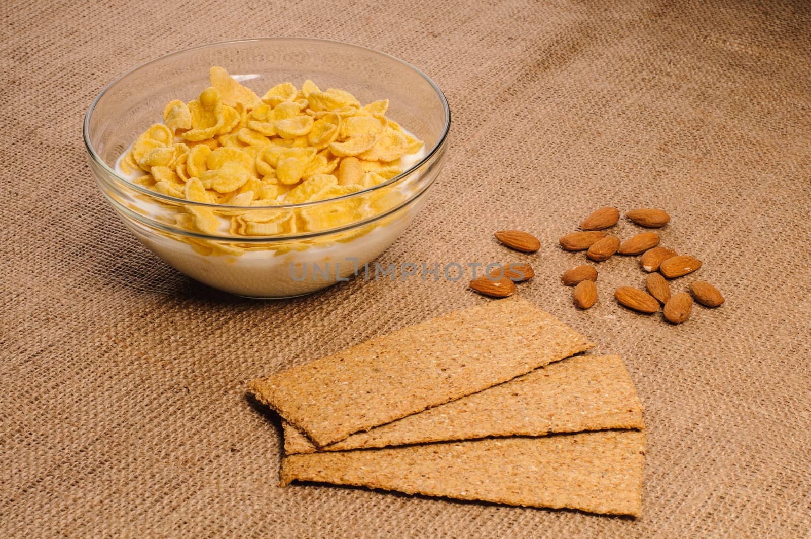 Bowl of cornflakes with milk, crispbreads and almonds on sacking background