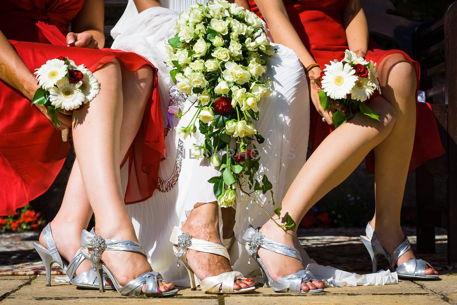 Feet of the bride among two bridesmaids sitting on chairs holding white flower bouquet in their lap and wearing beautiful shoes