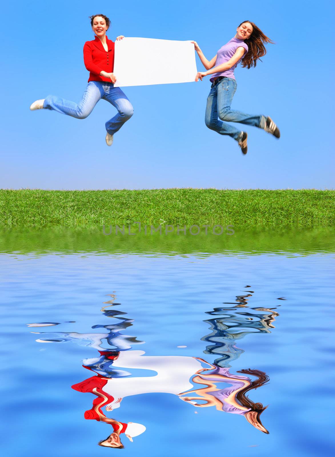 Girls jumping with blank sheet against blue sky with reflection on water