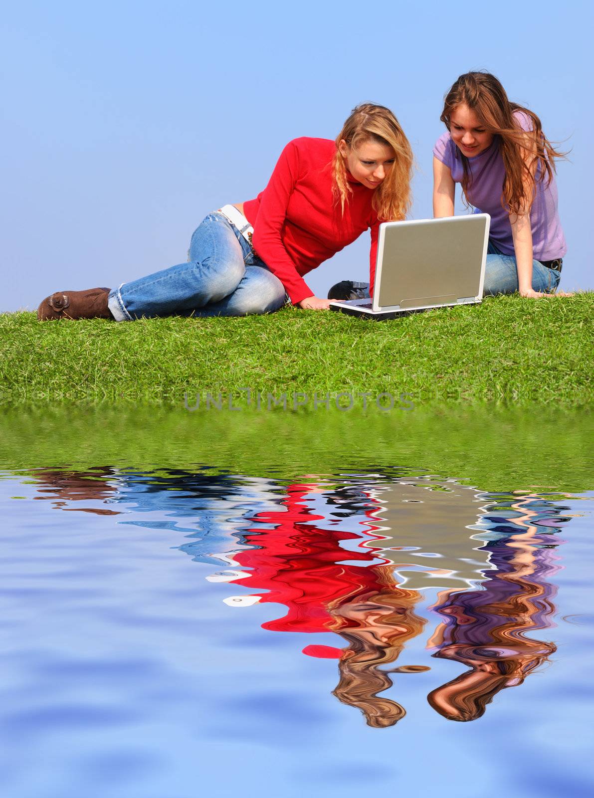 Girls with notebook sitting on grass against sky with reflection on water