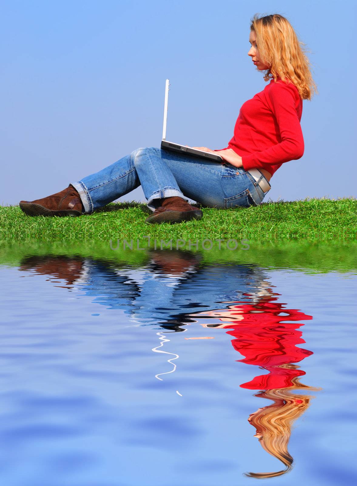 Girl with notebook sitting on grass against sky with reflection on water