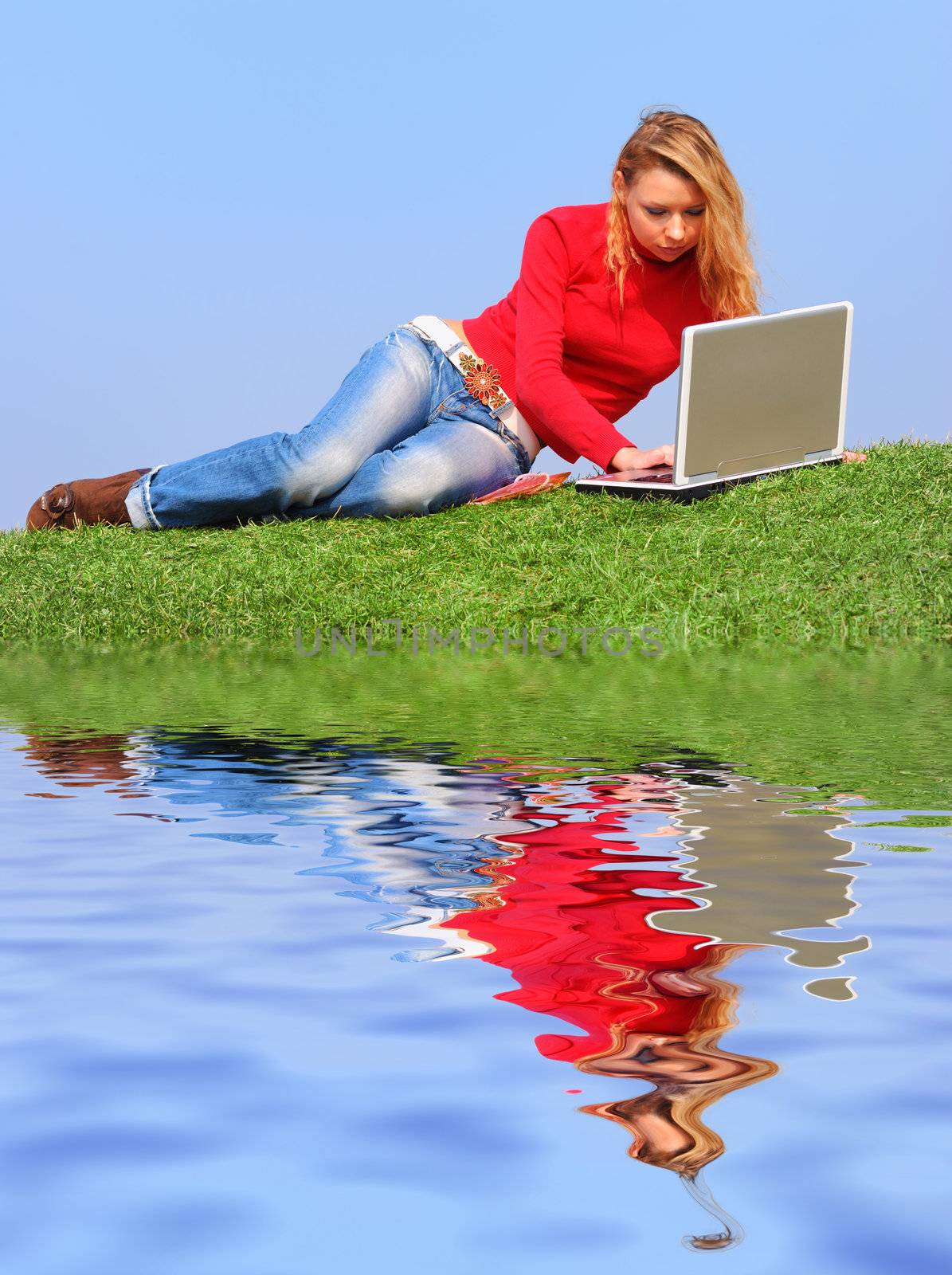 Girl with notebook sitting on grass against sky with reflection on water