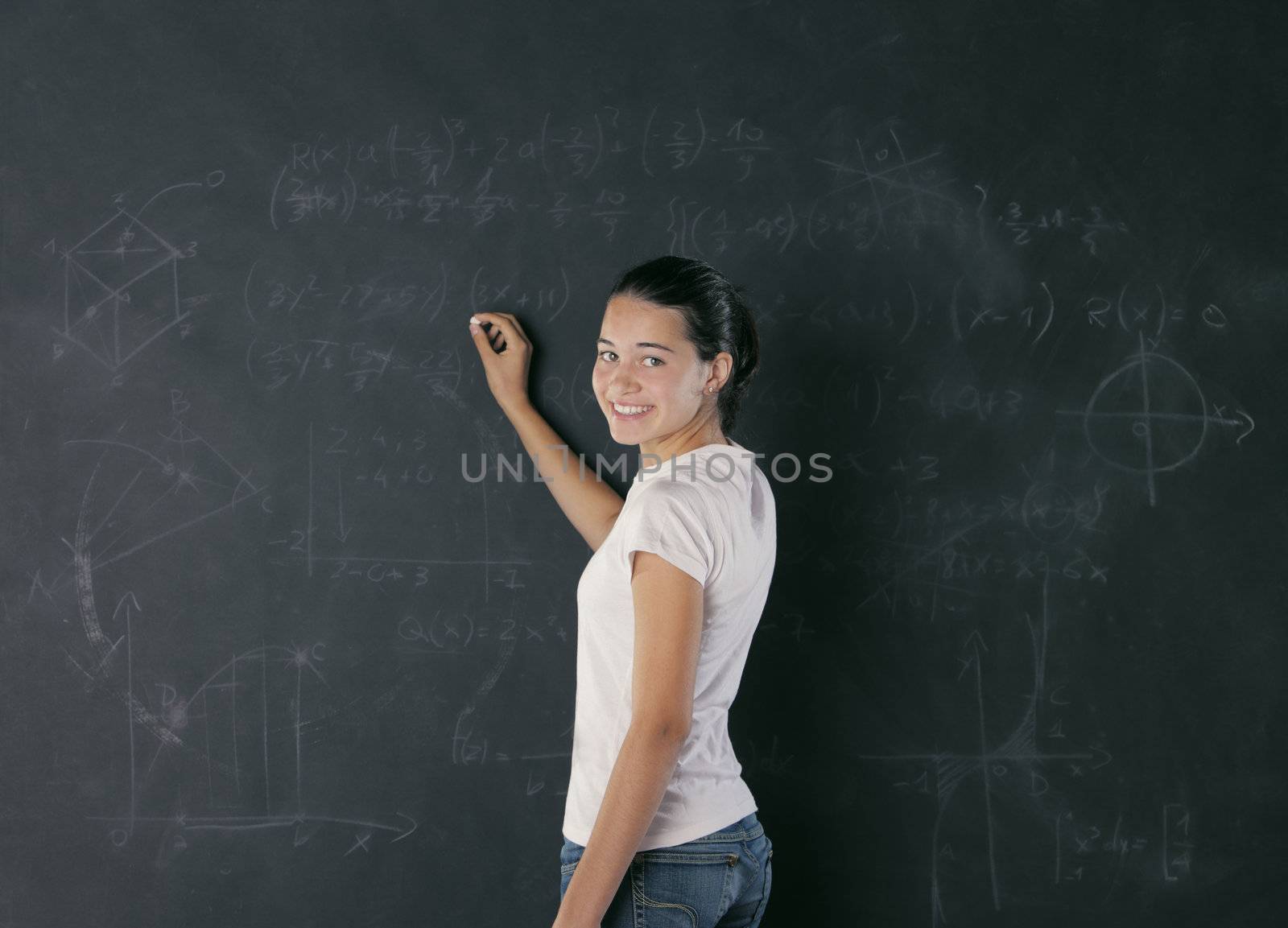 A young female student is standing in front of the blackboard going to draw something with a chalk.