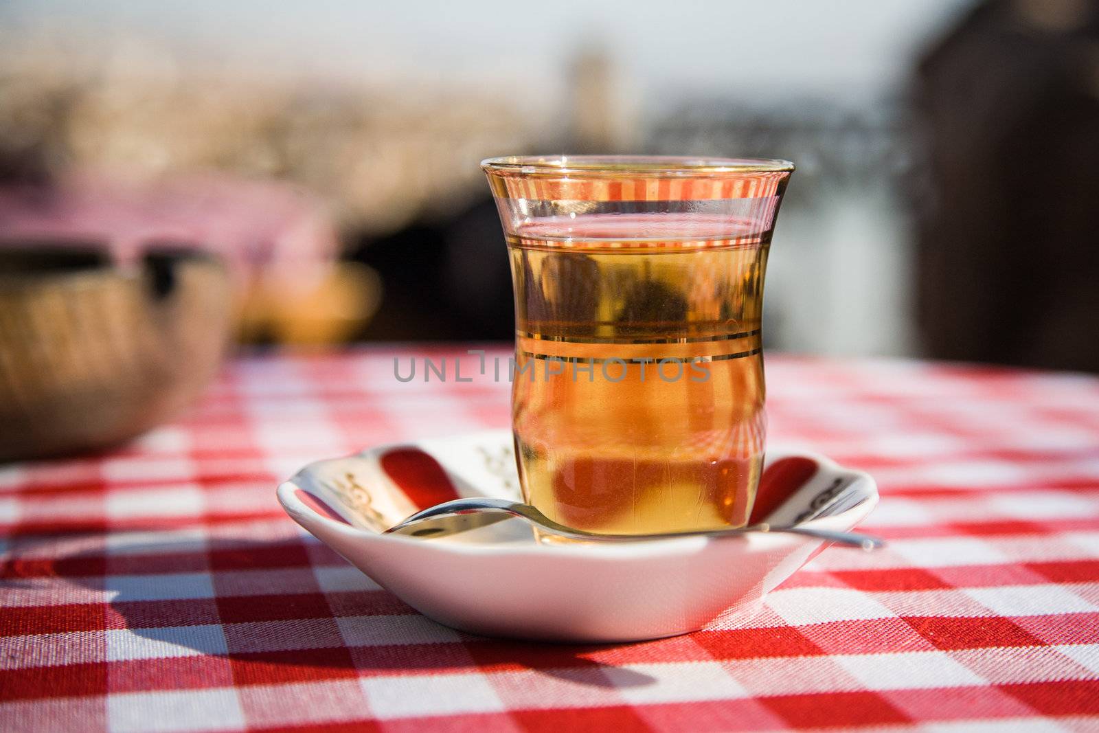Close up photo of a glass of turkish tea on saucer placed on a table which is covered by a red checked table cloth in a sunny day