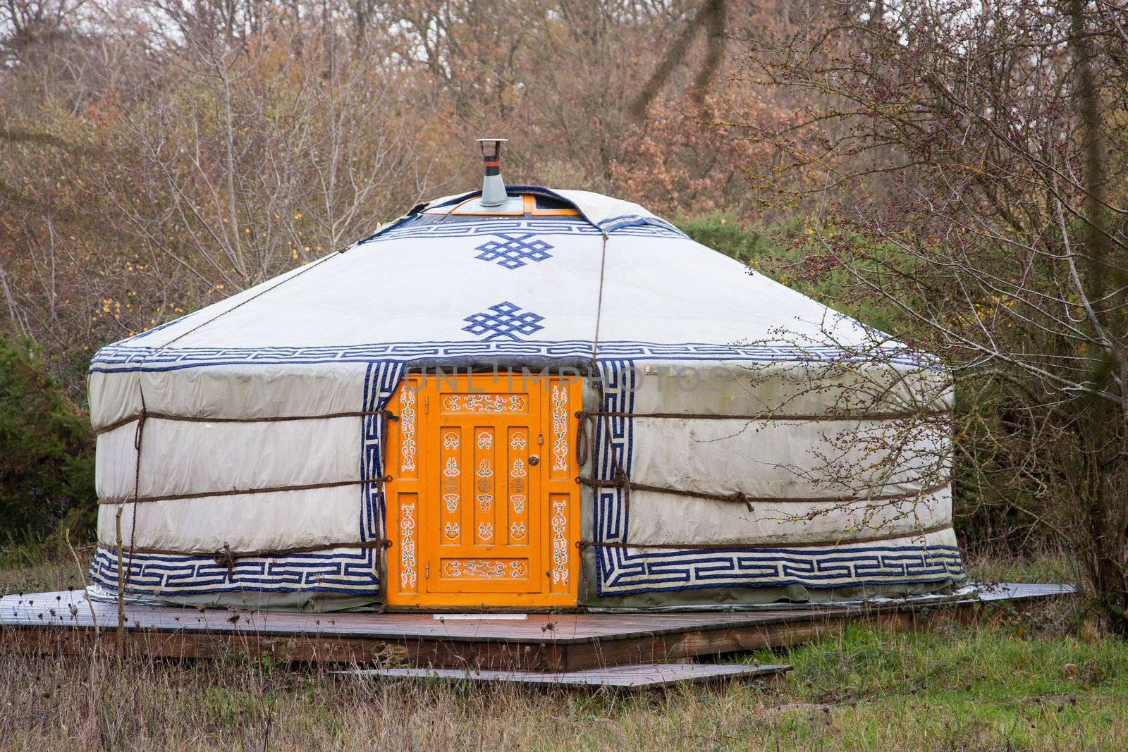Close-up photo of one white yurt with blue simbols and big closed well-decorated orange door in the forest on an autumn day