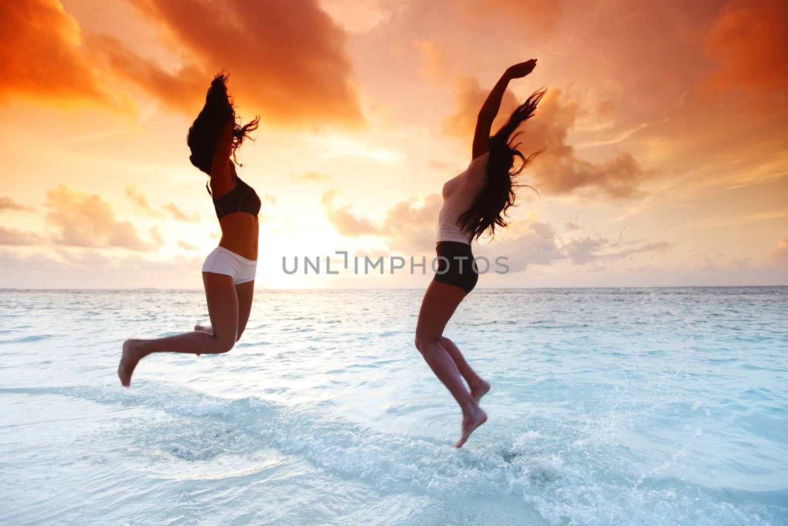 Two happy women jumping on sunset beach near sea