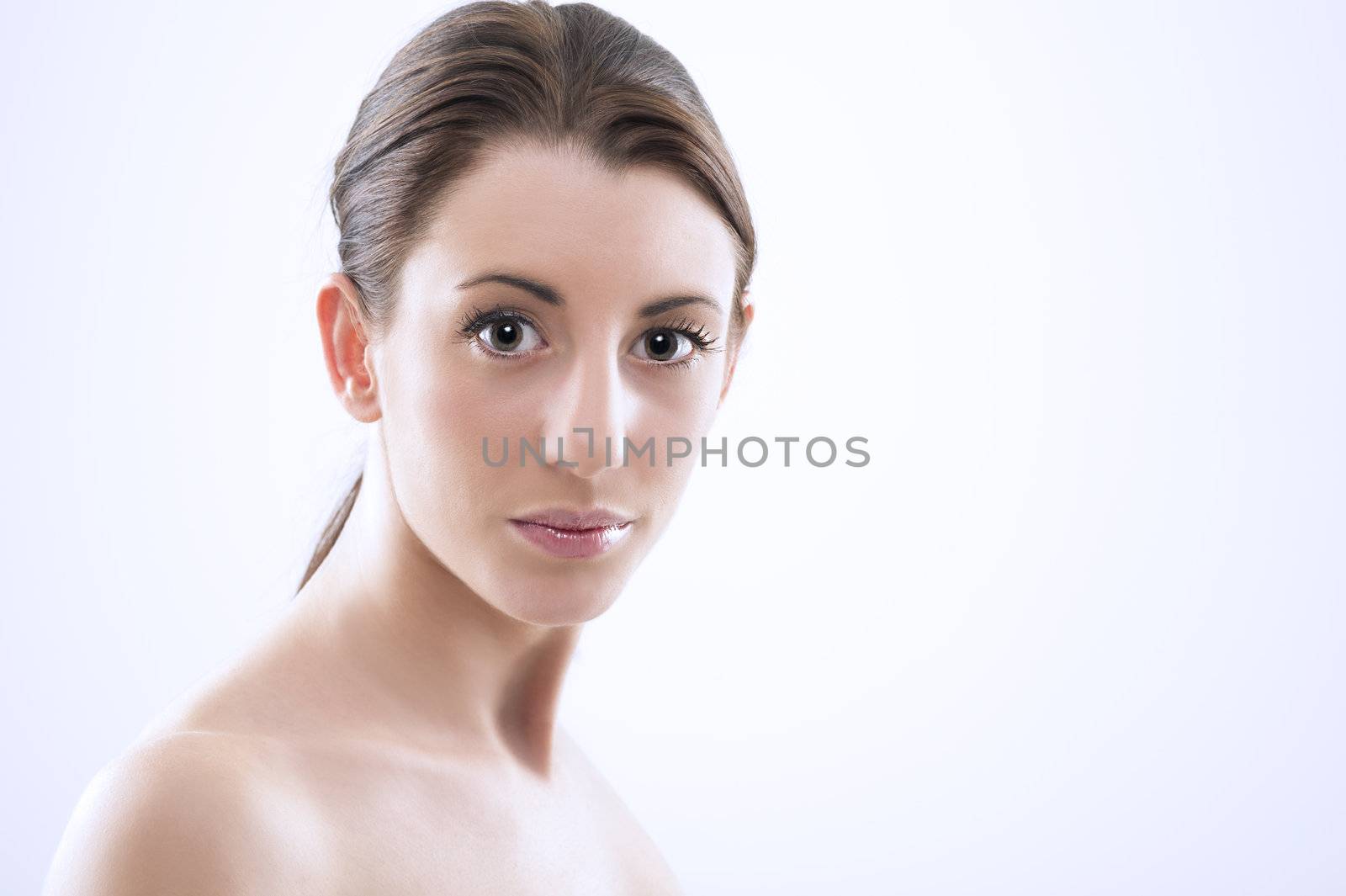 Head and shoulders studio portrait of a beautiful sexy woman with bare shoulders, a natural complexion and her hair tied back in a ponytail looking at the camera with large lustrous eyes