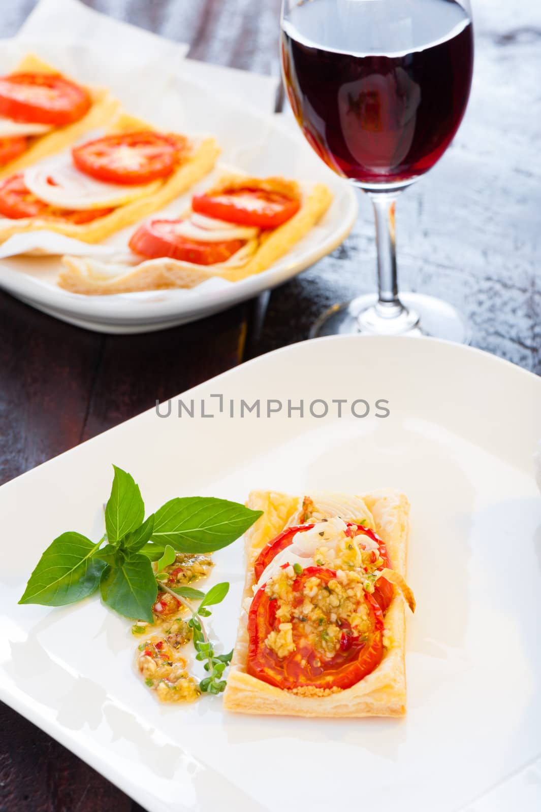 A small piece of mini pizza with some herbs on white plate and a glass of red wine in the background